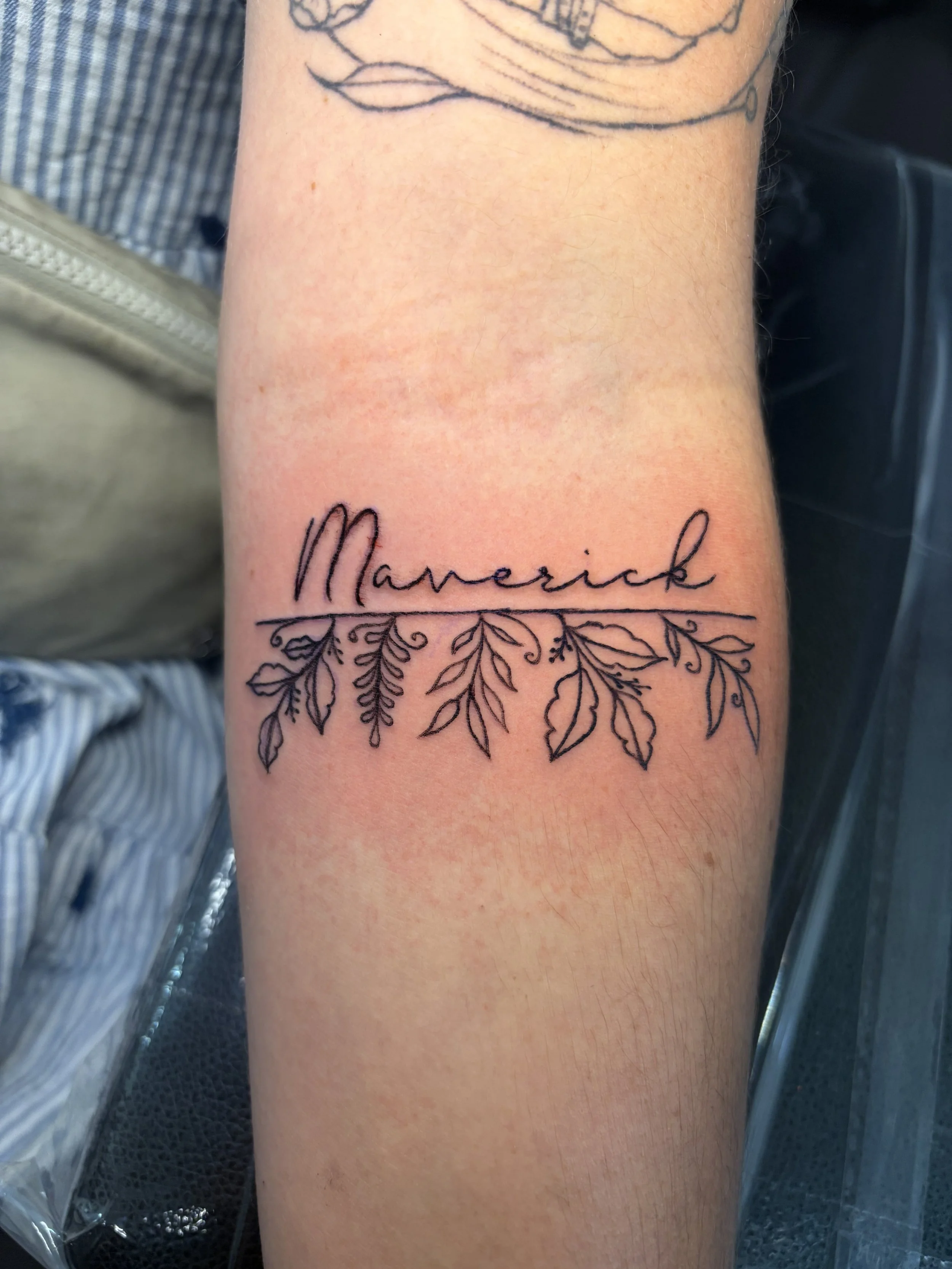 A tattoo on a person's forearm featuring the word ' Maverick' written in cursive, with decorative floral and leaf designs surrounding it.