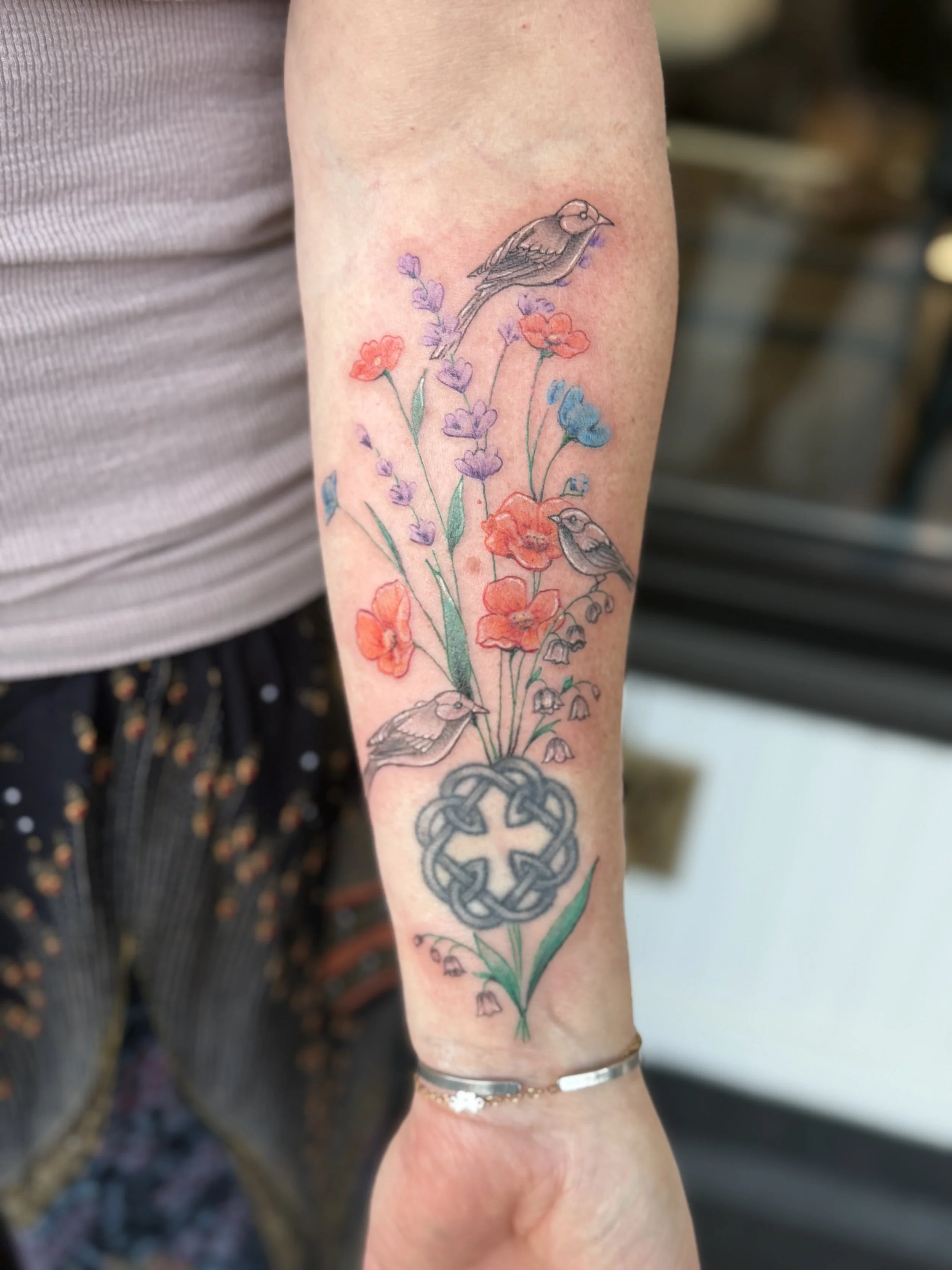 A tattoo of three small birds perching on a colorful floral arrangement on a person's forearm, with a Celtic knot at the wrist.
