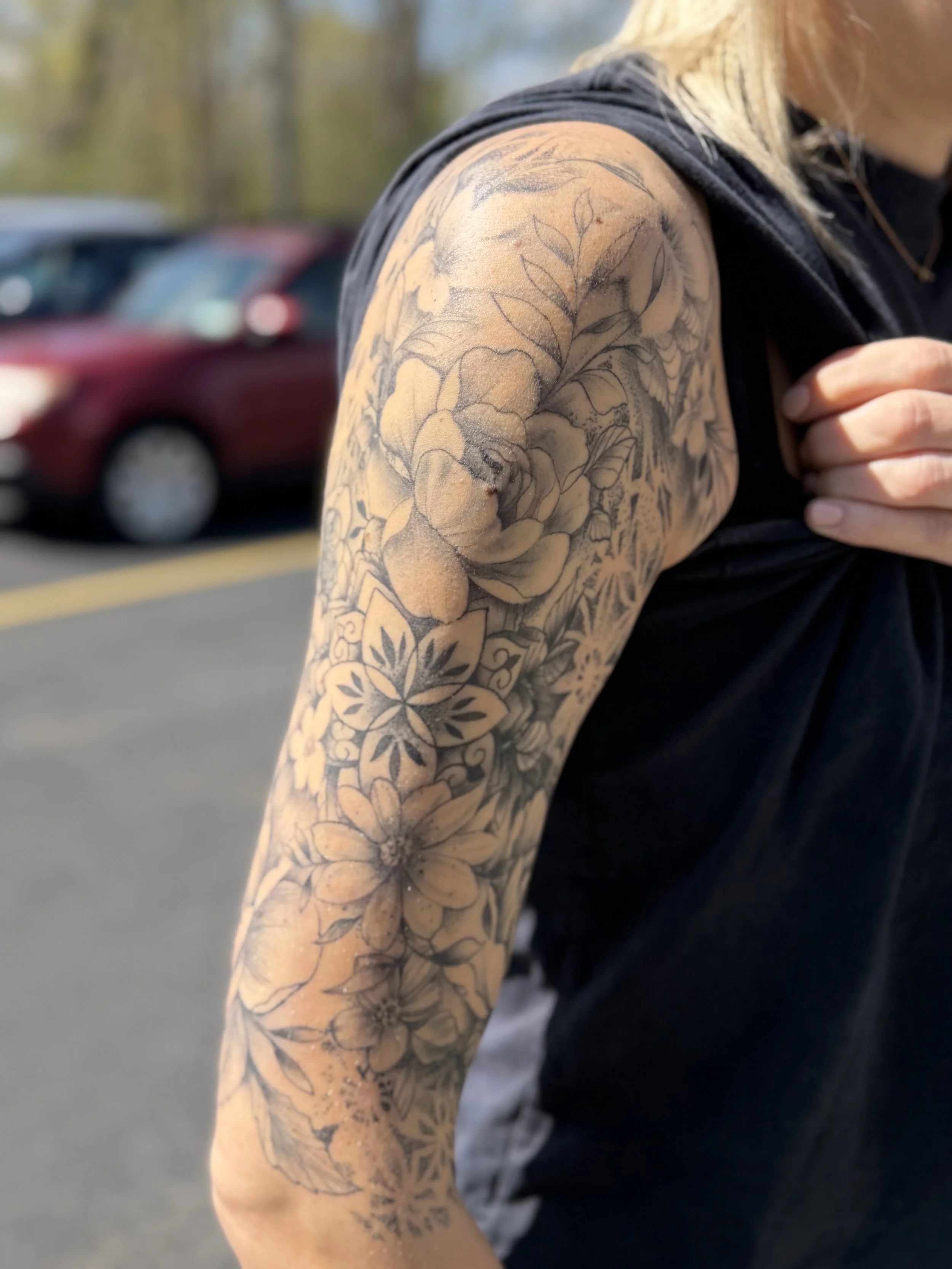 Close-up of a person's arm with a detailed black and gray floral tattoo, wearing a sleeveless black top in an outdoor setting.