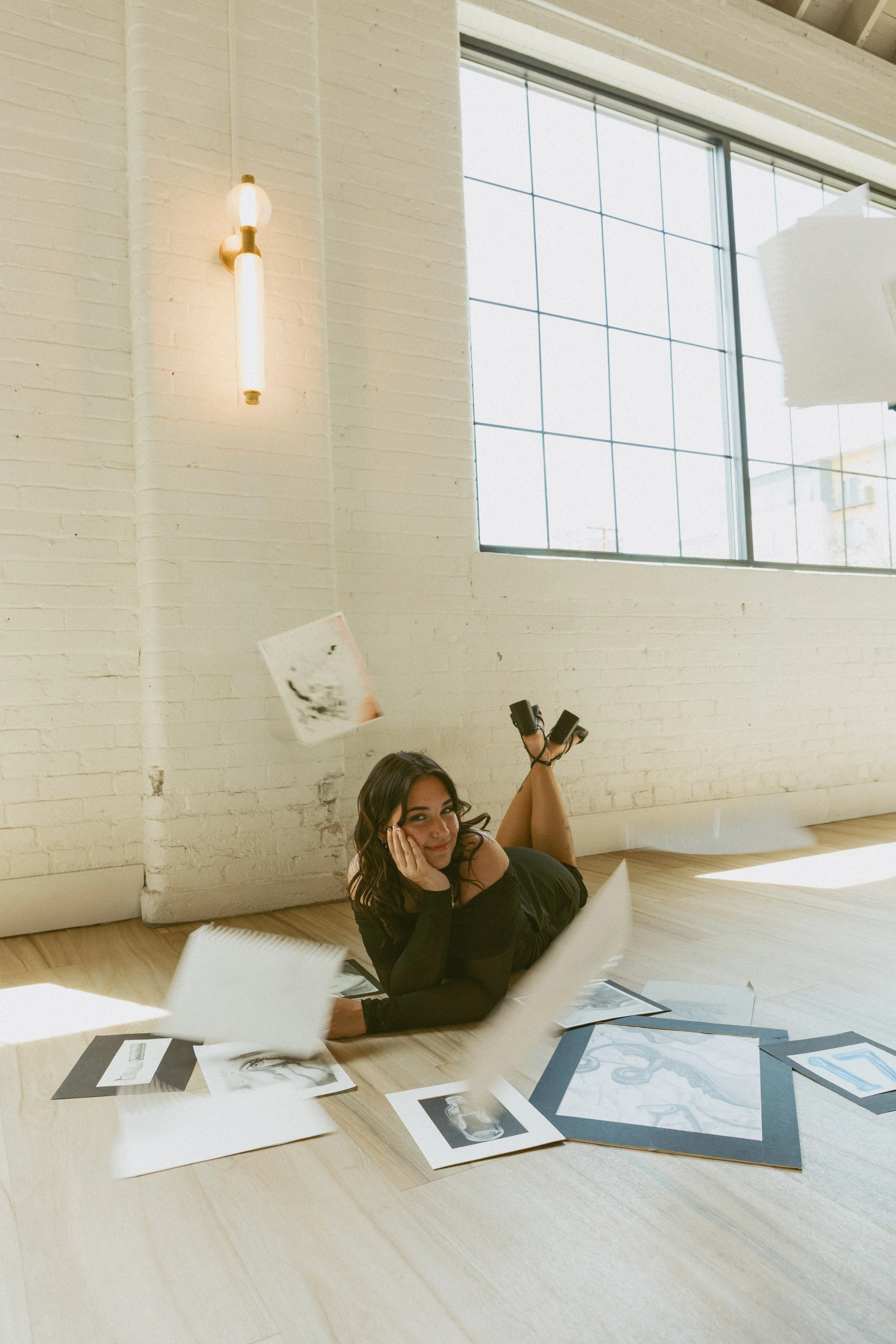 A woman lying on the floor surrounded by scattered art prints and sketches, smiling while holding her face in her hand, in a bright room with large windows and white brick walls.