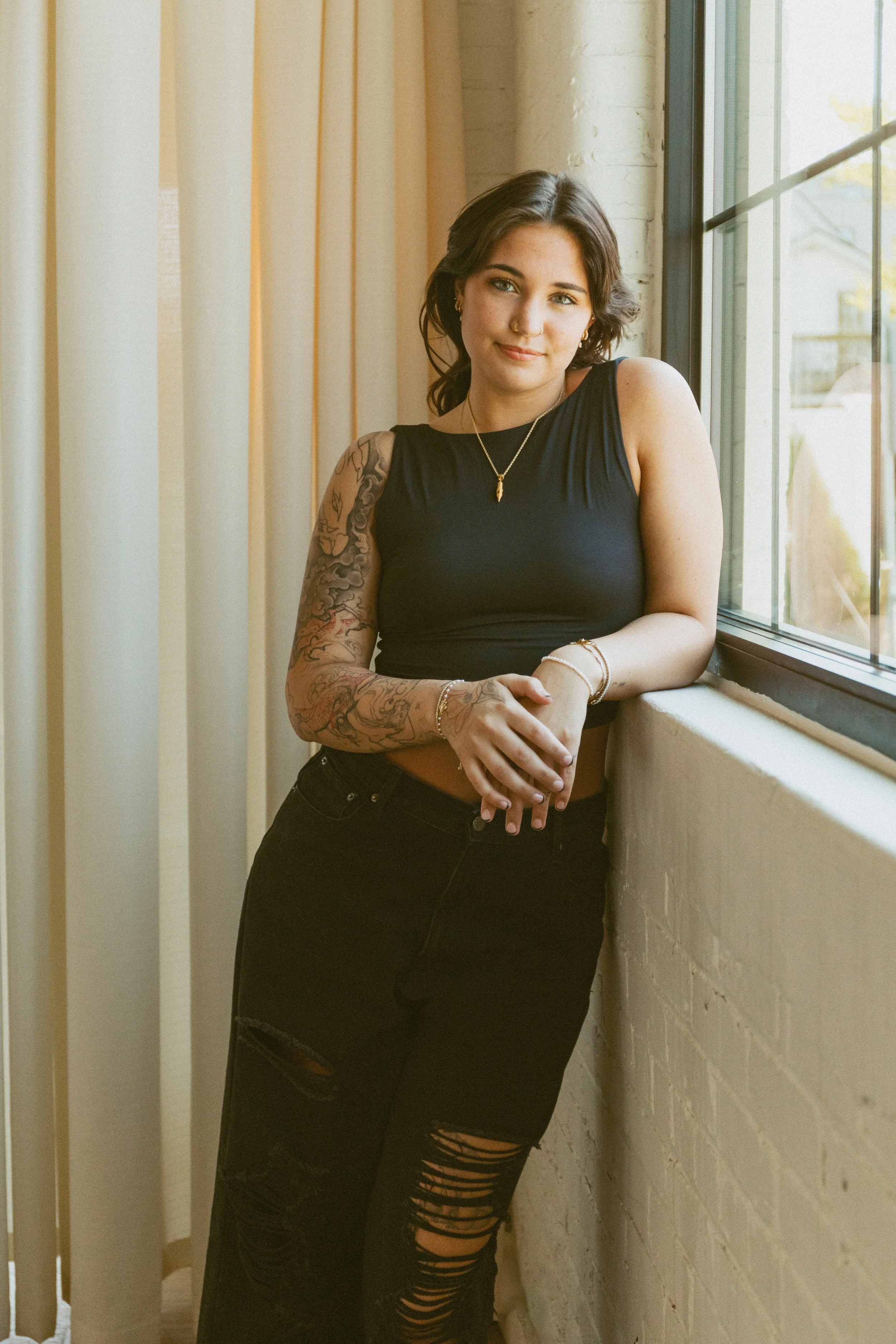 A young woman with dark hair and tattoos is standing by a window, wearing a black sleeveless top and ripped black jeans, with jewelry including earrings, necklaces, and bracelets.