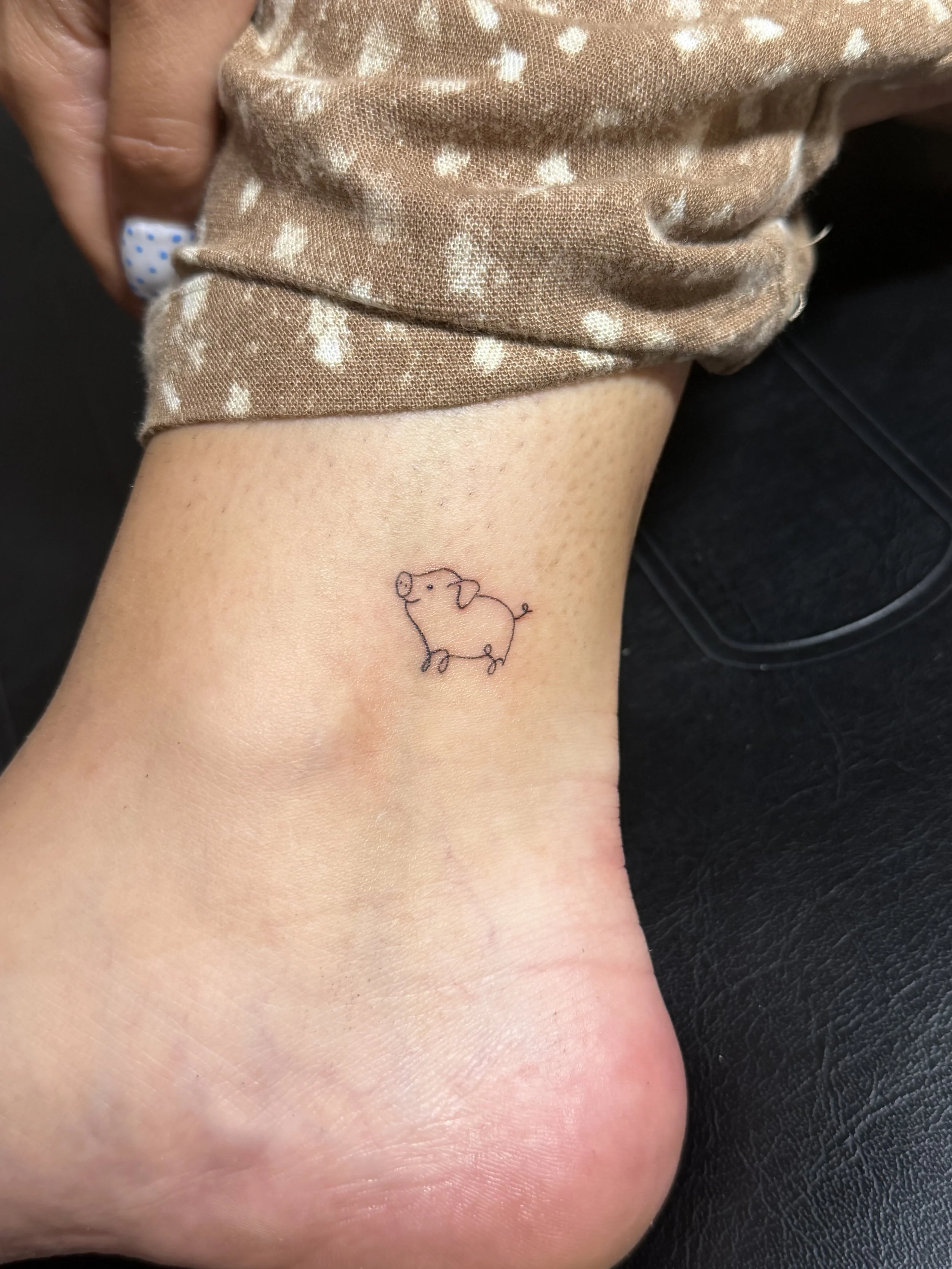 A tattoo of a minimalist line drawing of a pig is on a person's ankle, with the person wearing a brown polka dot fabric.