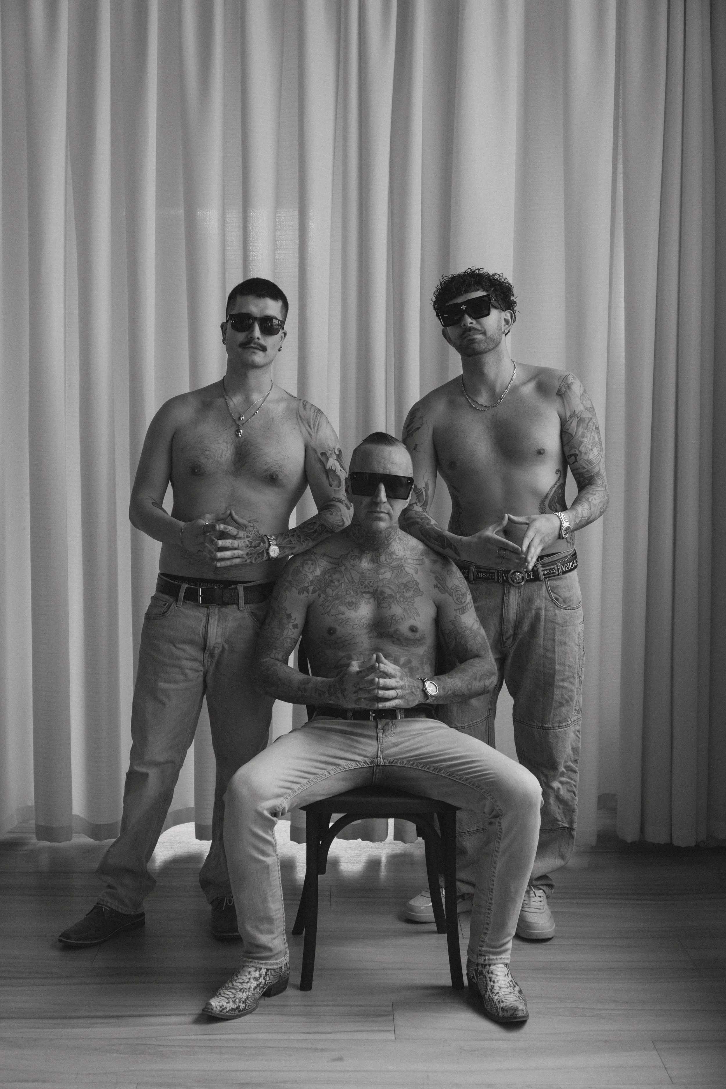 Four shirtless men with tattoos and sunglasses posing in front of a curtain. One seated man with tattoos and snake skin-patterned boots in front. Two standing men with tattoos and chain necklaces behind, one in jeans with a Versace belt, the other wi