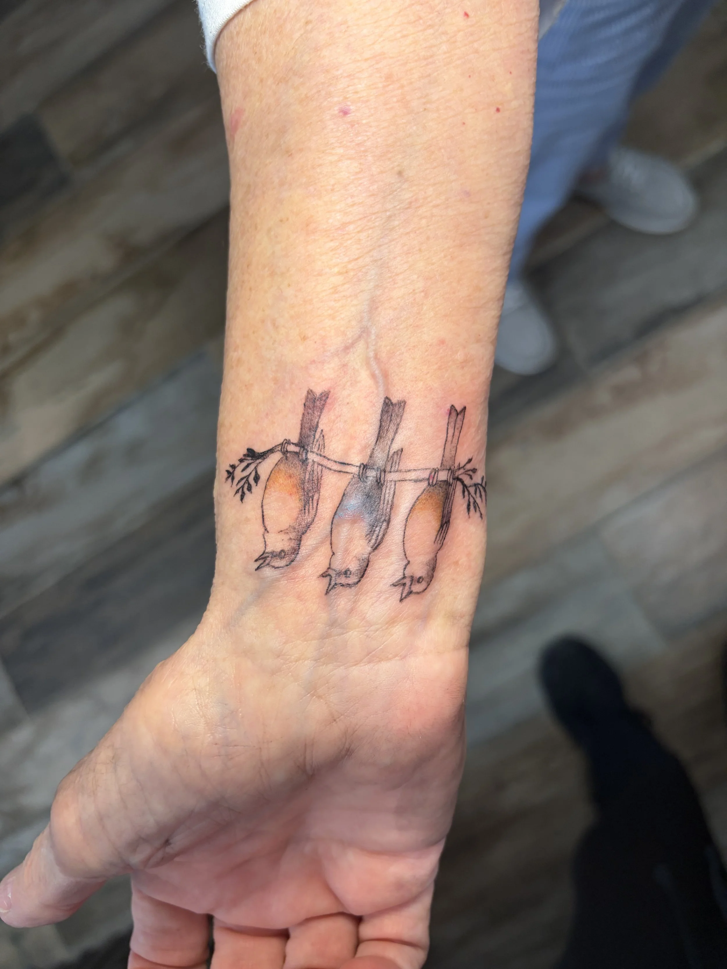 A person's arm with a tattoo of three birds perched on a barbed wire. The background appears to be a wooden surface.