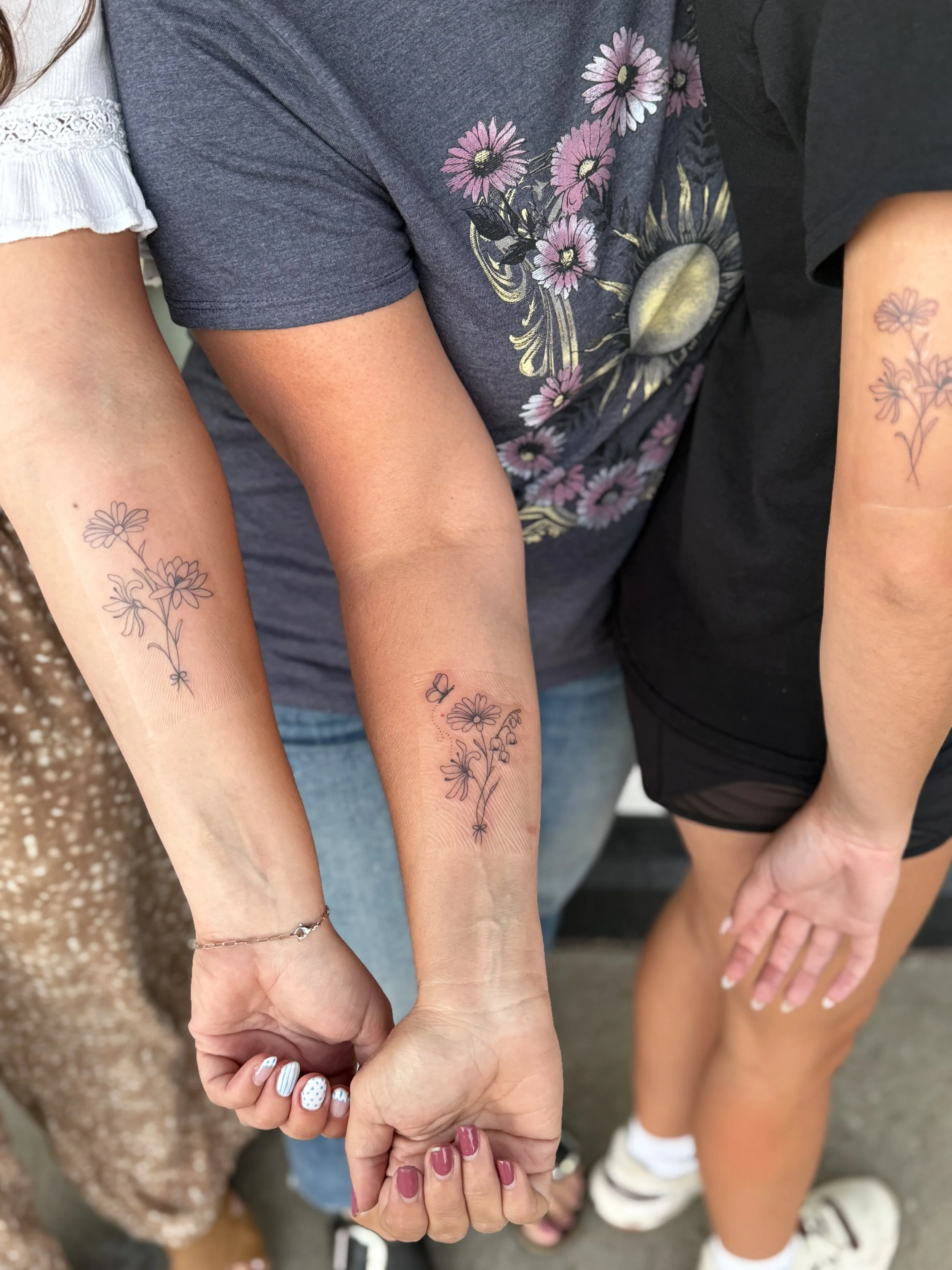 Multiple people holding hands, showing tattoos of flowers and butterflies on their arms, with colorful nail polish and casual clothing.
