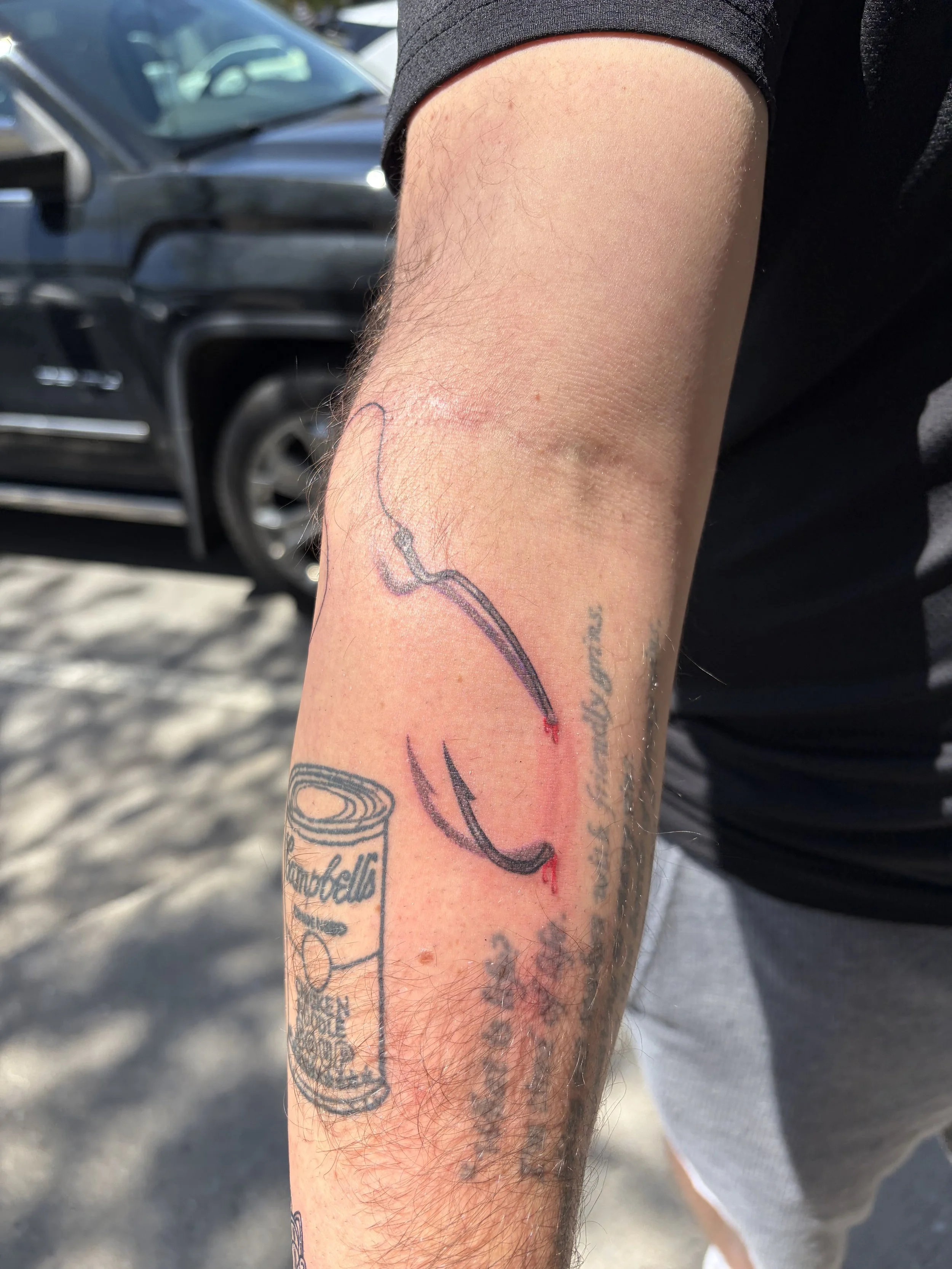Close-up of a person's arm showing fresh tattoos, including an outline of a wine glass with the word 'Countess' and some other script. The arm is slightly hairy and the background shows parked cars and an outdoor setting.
