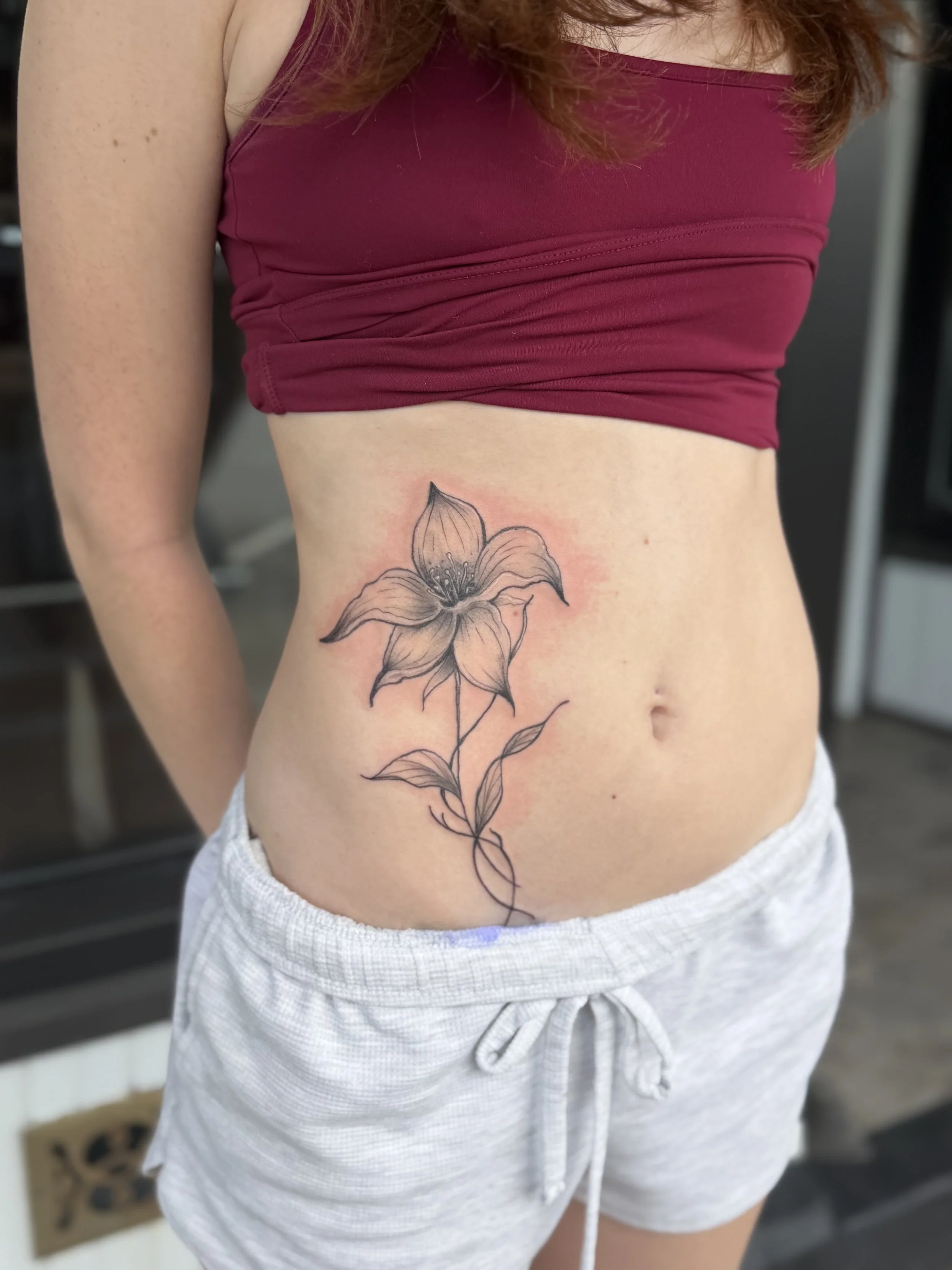 Close-up of a woman wearing a maroon crop top and gray shorts, showing a detailed black and gray floral tattoo on her lower left side near her waist.