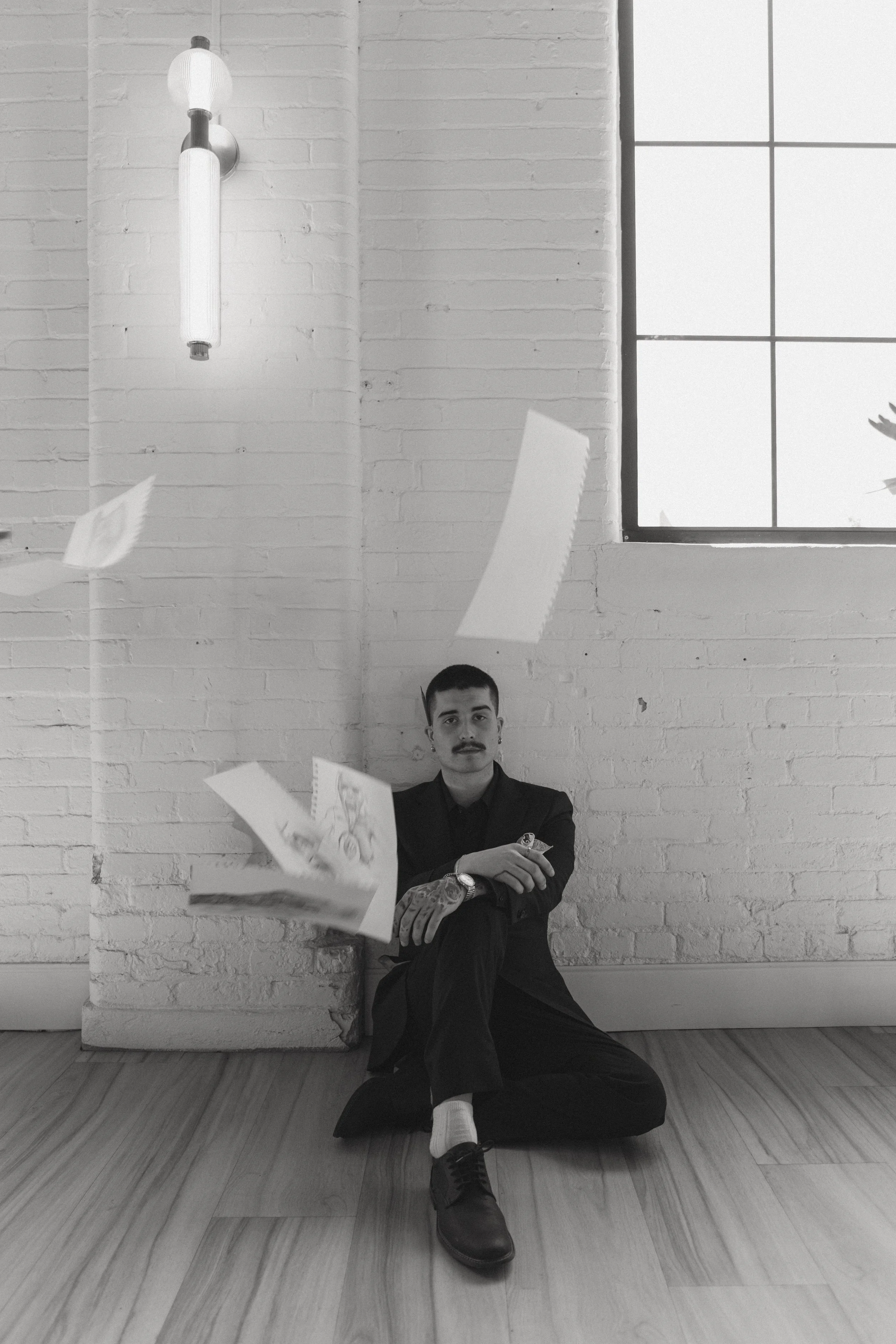 A man with a mustache and tattoos, dressed in black, sitting on the wooden floor with crossed legs in front of a white brick wall and large window. Papers are flying in the air around him.