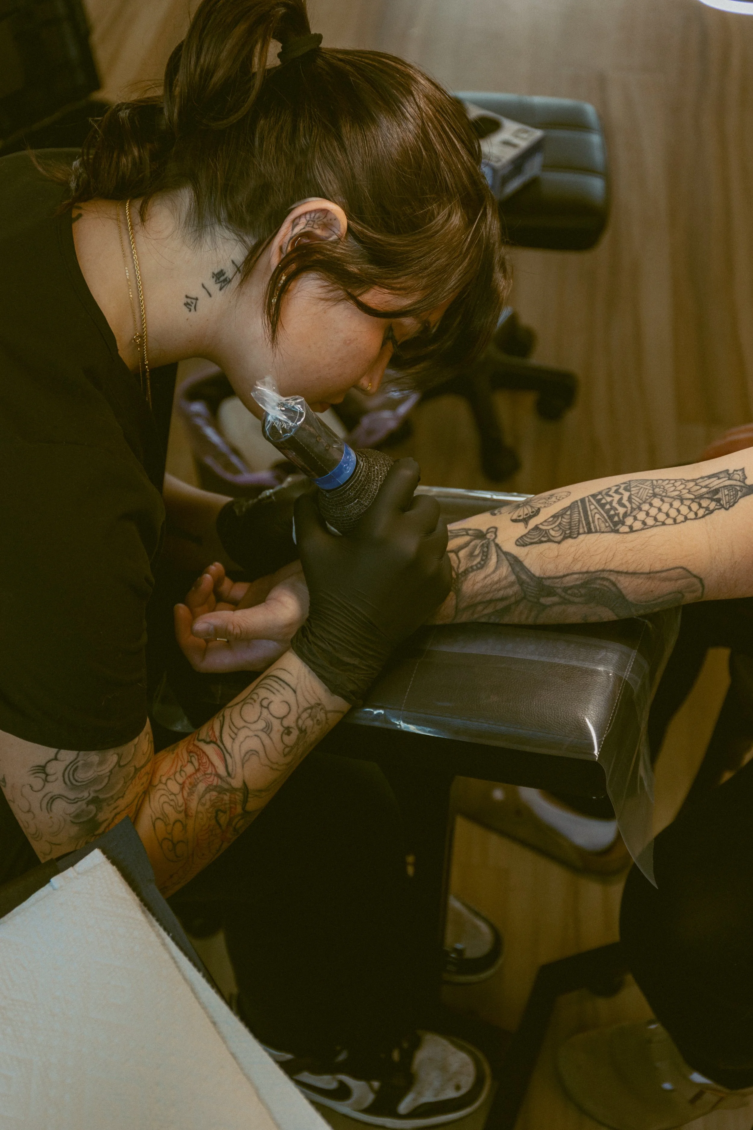 A person with tattoos on their arms and neck is getting a tattoo on their leg by a tattoo artist. The person is seated, and the artist is using a tattoo machine. The person has brown hair tied up and is wearing black gloves.