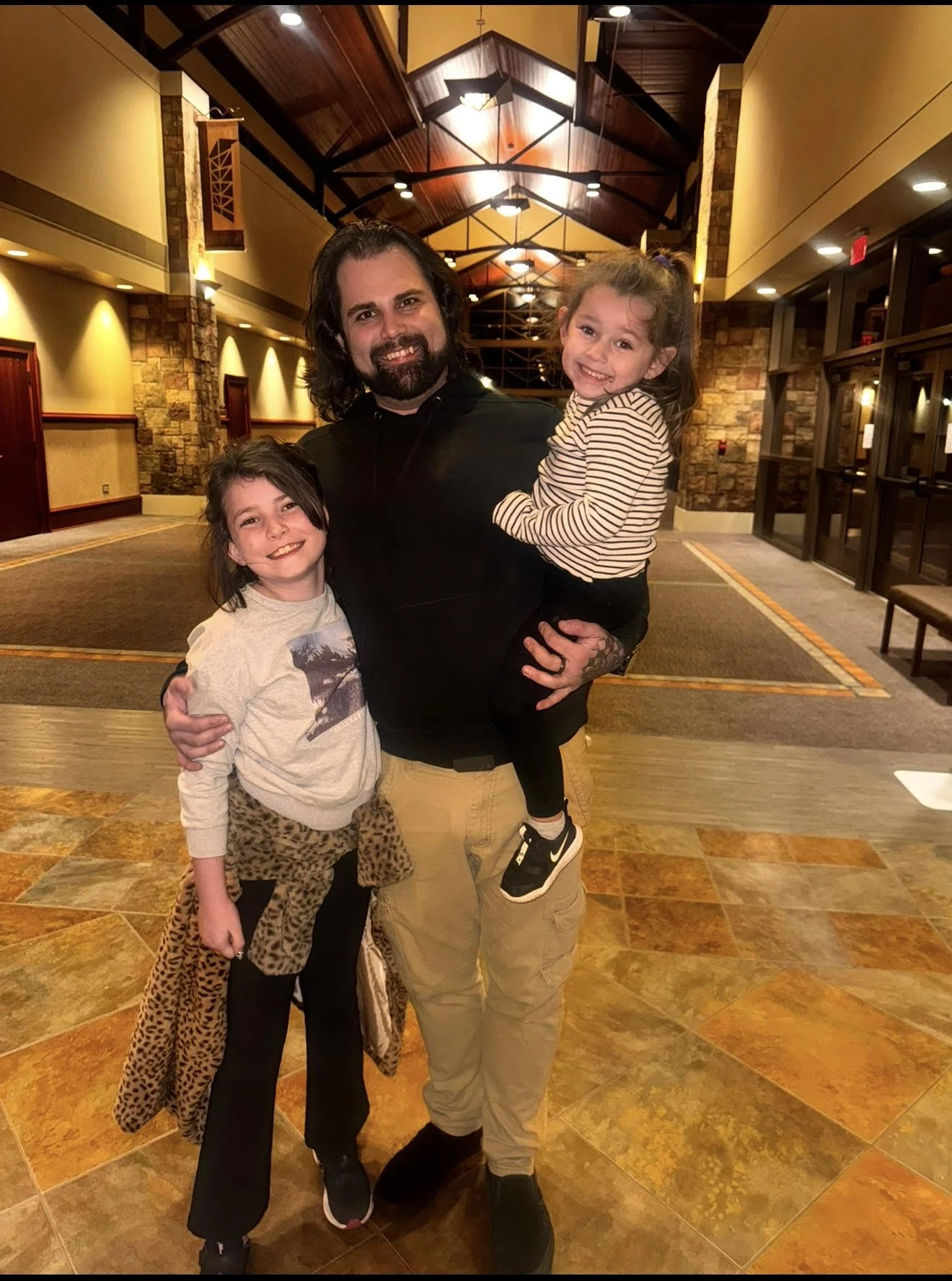 A man holding a young girl in his arms and standing next to another girl in a lobby with wooden and stone decor.