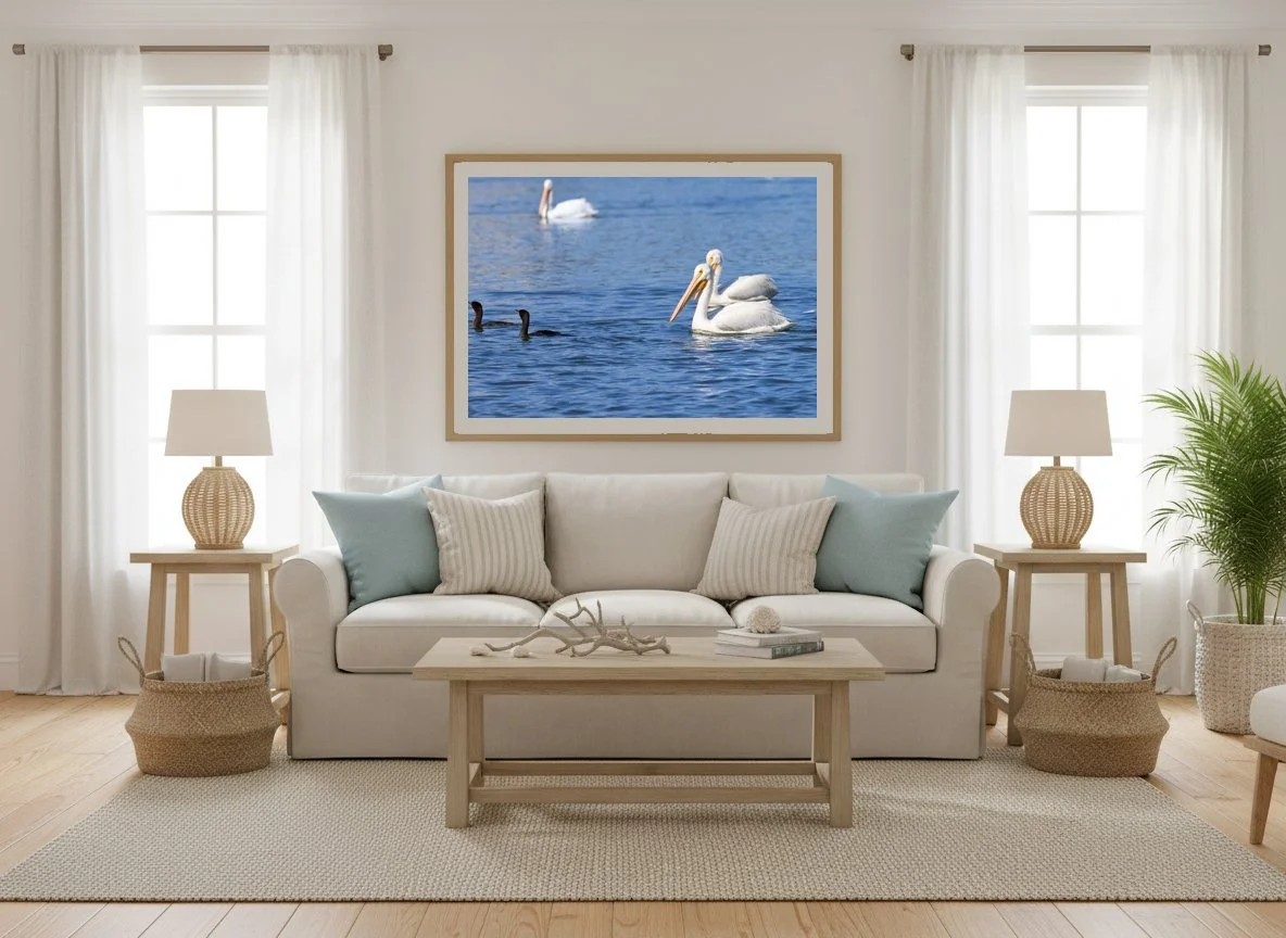 Living room with white sofa, light blue and beige pillows, wooden coffee table, two side tables with lamps, a framed photo of ducks and swans on the wall, a plant in a white pot, and baskets on the floor.