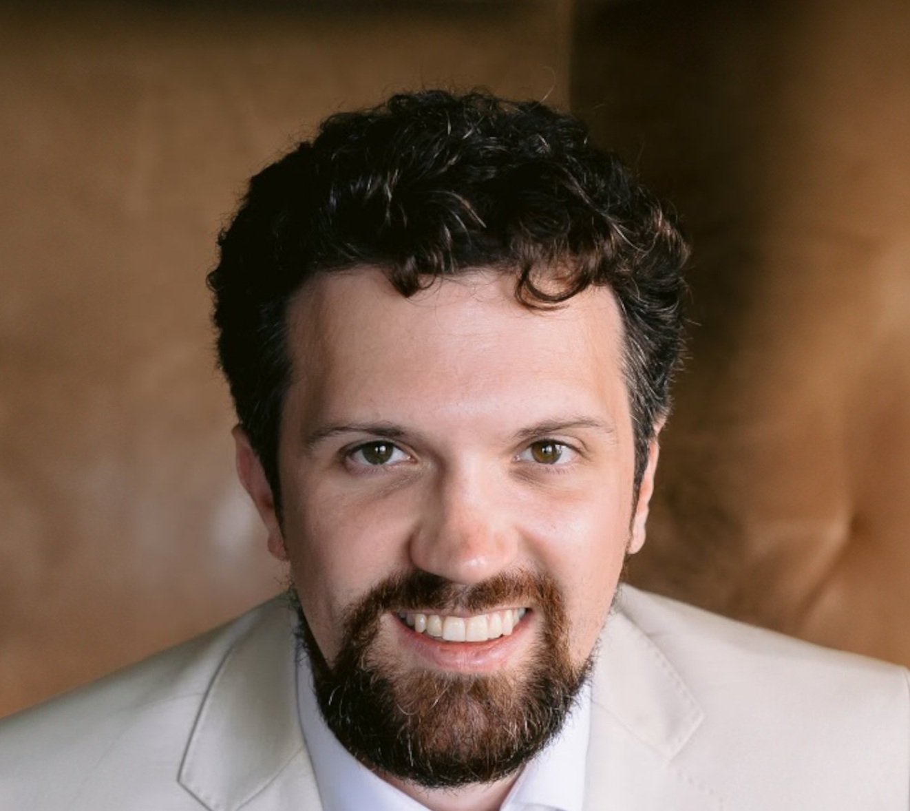 Headshot of John O'Brien. Smiling man with curly hair and beard in a light suit, brown background.