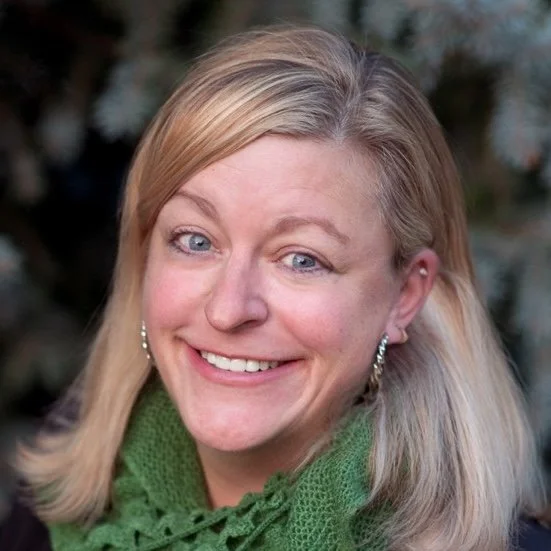 Smiling person with blonde hair, wearing green scarf and earrings, outdoors.
