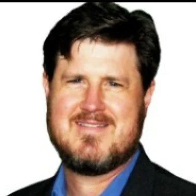 Smiling man with a beard and dark hair wearing a blue shirt and dark blazer, white background.
