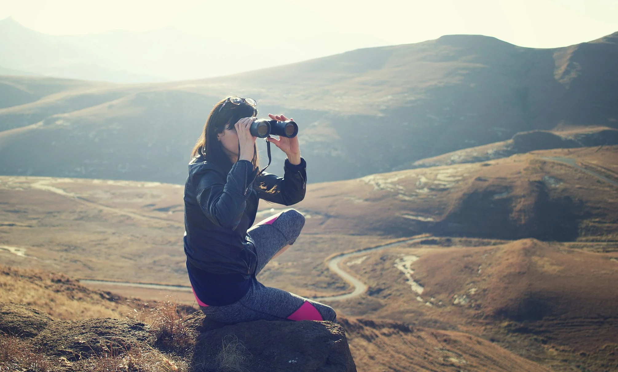 A woman sitting on small mountain as part of a hike using binoculars