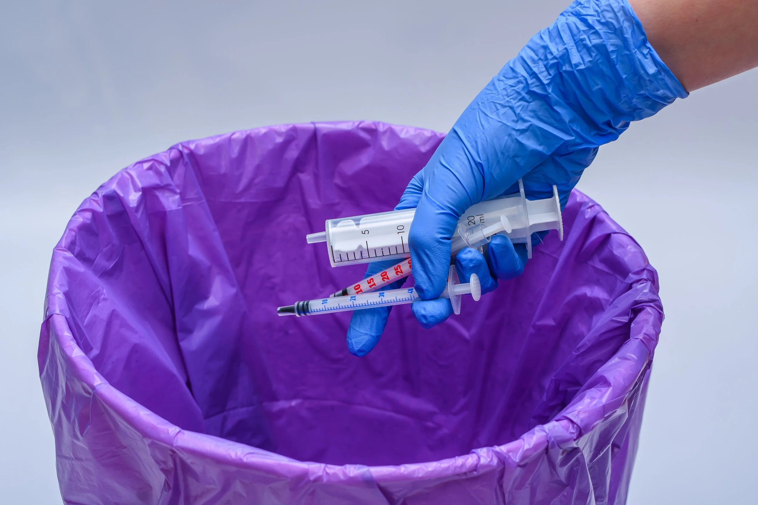 An image of a latex gloved hand throwing away multiple syringes into a trash can