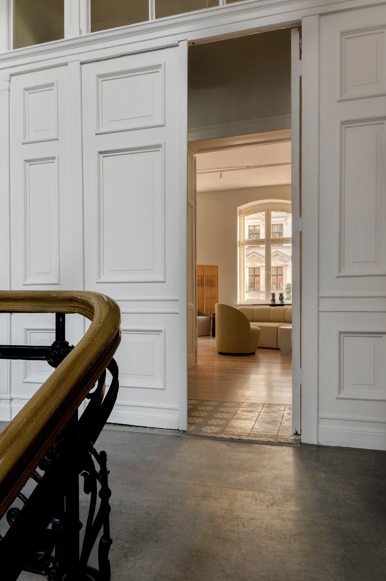 View of an interior space with a staircase leading up to a white doorframe that opens into a room with a large window and seating area.