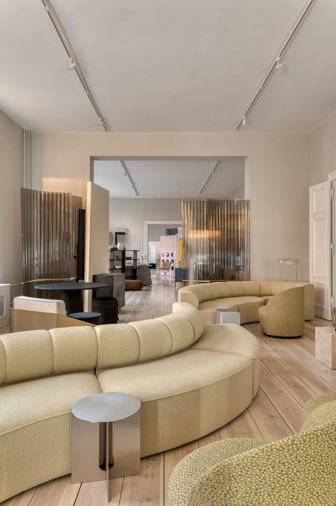Modern living room with beige curved sectional sofas, a small side table, patterned green chairs, wood flooring, and decorative room dividers.