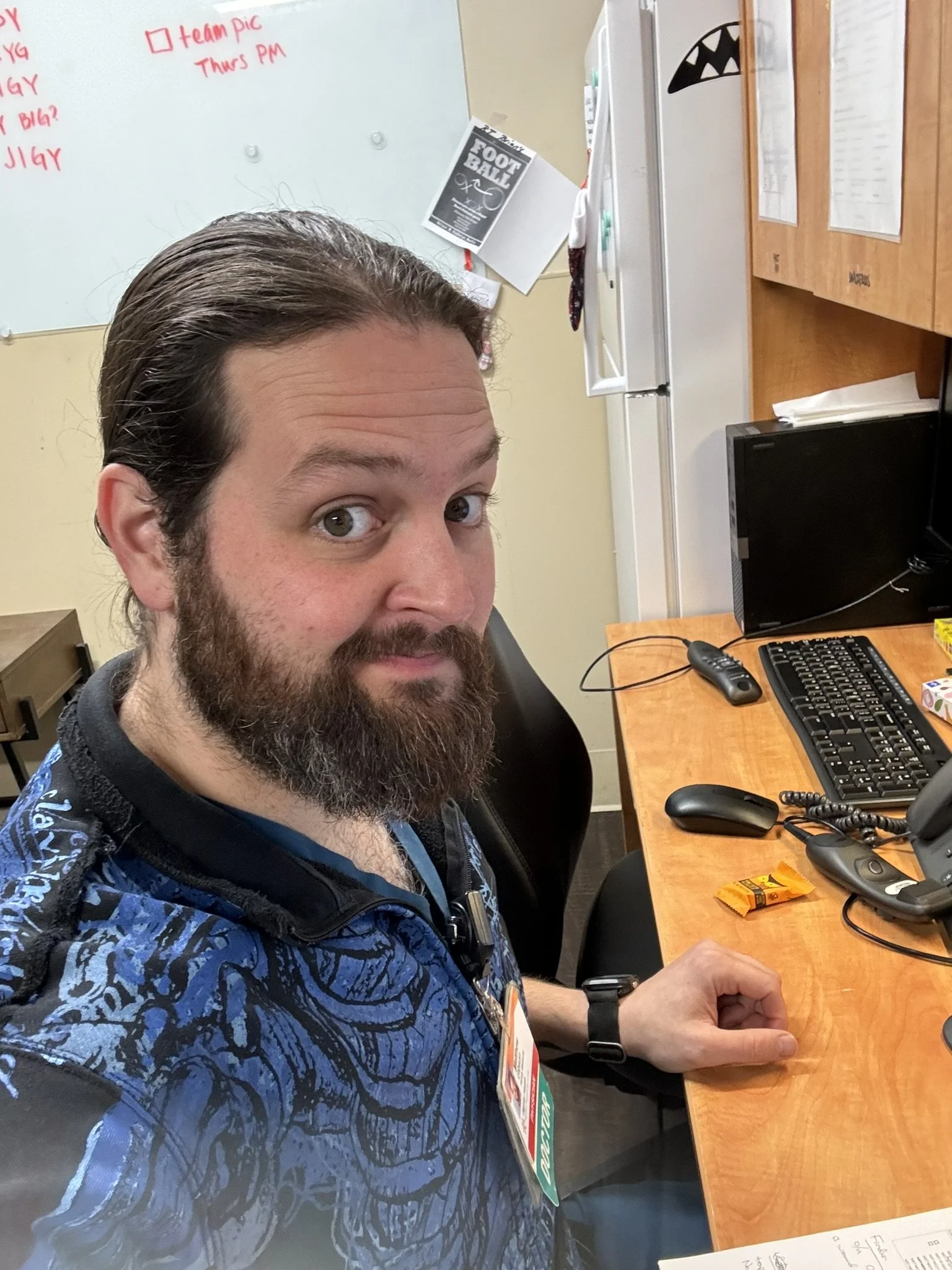 A man with a beard and long hair taking a selfie at a desk with a computer monitor, keyboard, mouse, and a snack. Behind him is a whiteboard and cabinets.