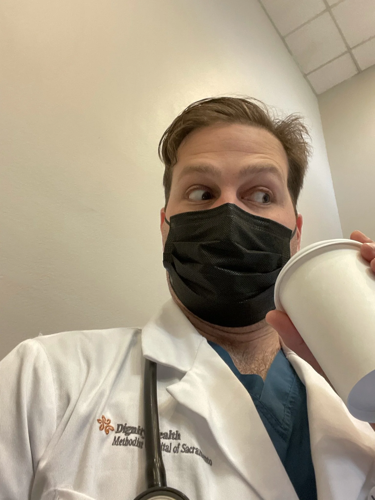 A healthcare worker in scrubs and a black face mask holding a white disposable cup, standing against a plain wall with a tiled ceiling.