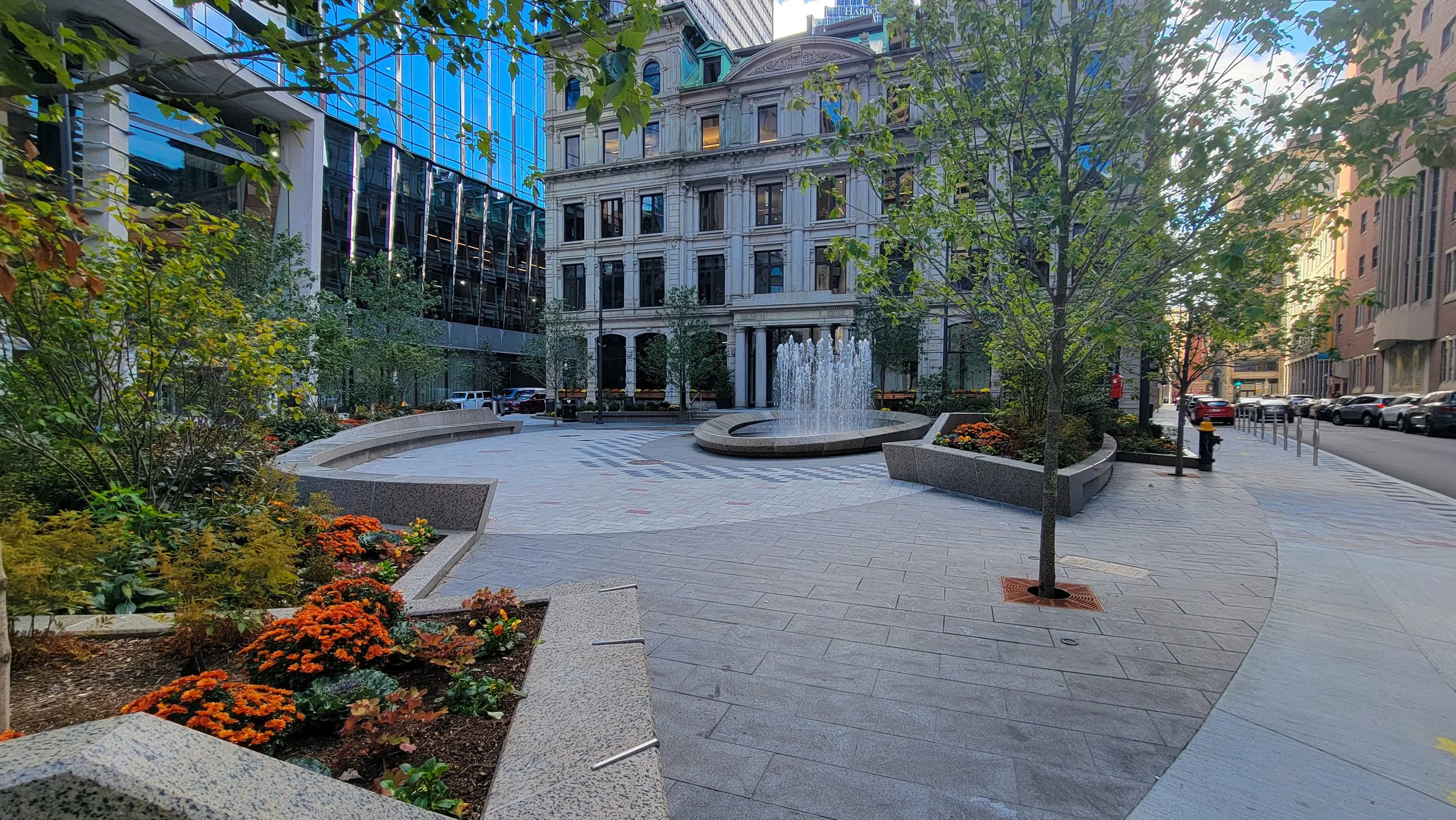A city plaza with trees, flower beds, a fountain, and surrounding tall modern buildings.