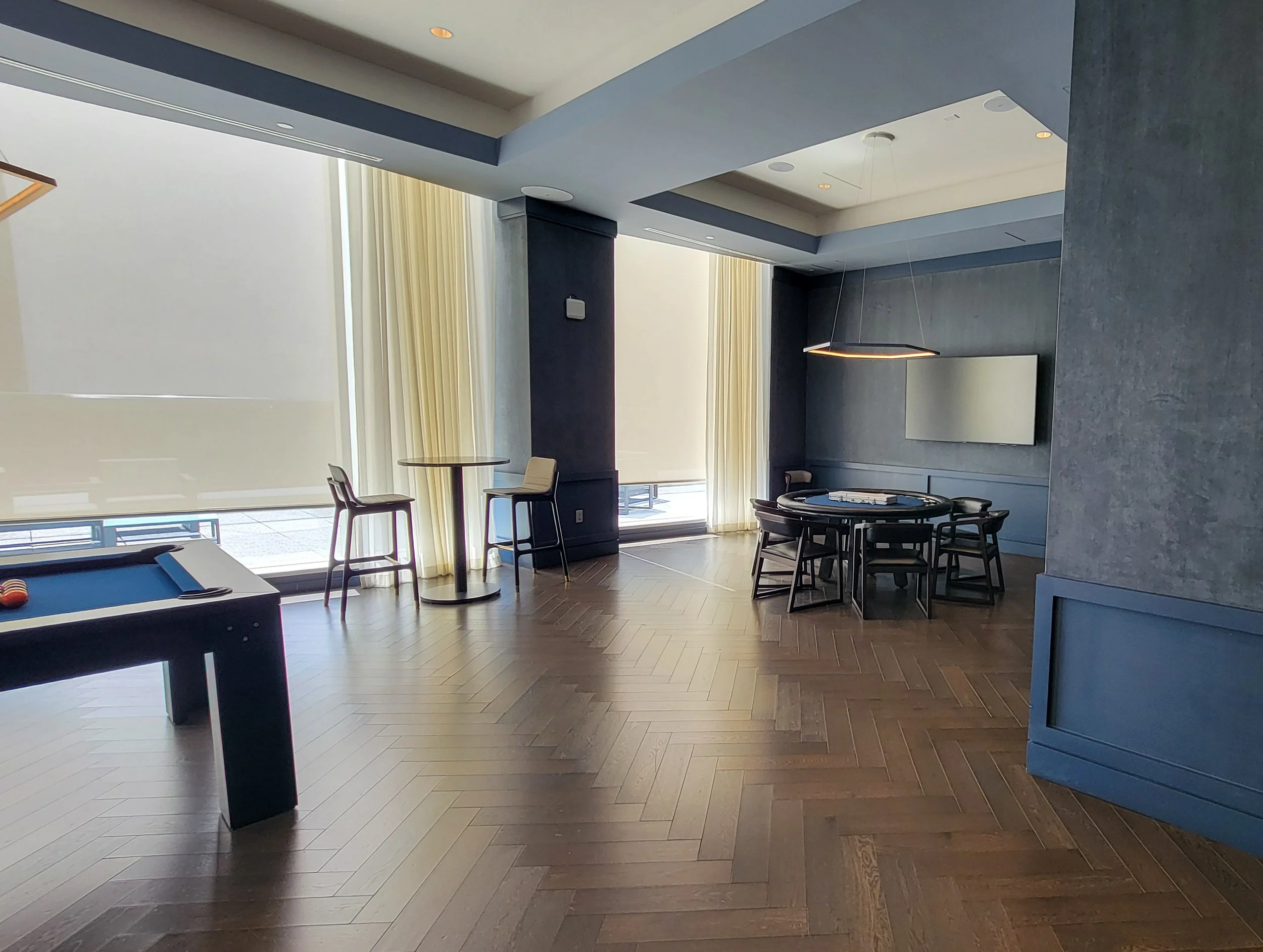 Interior of a modern lounge with a pool table, a round table with chairs, large windows with light curtains, and a wall-mounted TV.