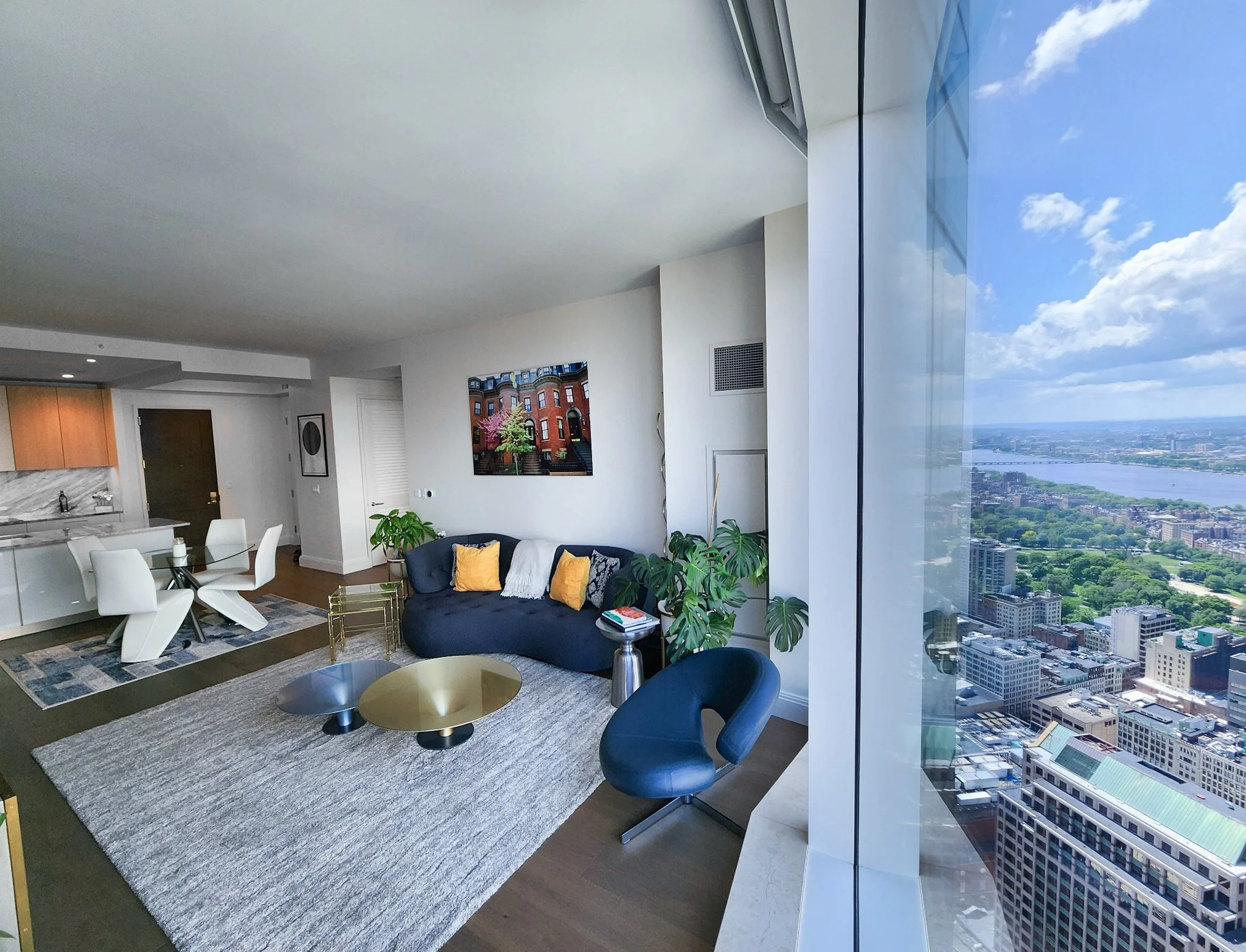 Modern high-rise apartment living room with large floor-to-ceiling window overlooking a cityscape and river, featuring a navy sofa with yellow and white pillows, a black leather chair, round coffee tables, and green potted plants.
