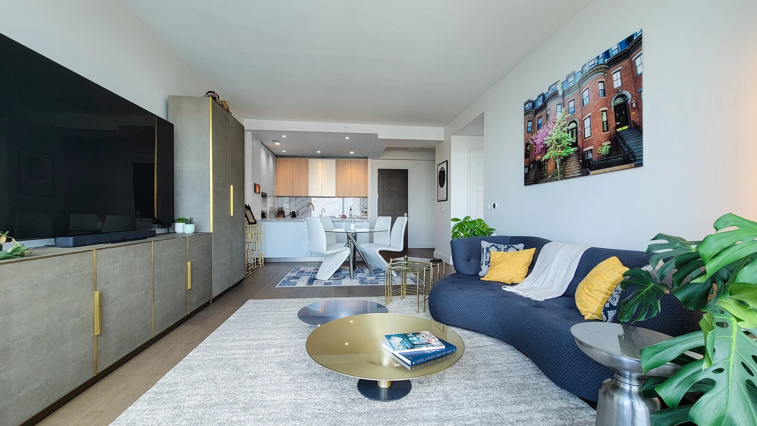 Living room with dark blue couch, yellow cushions, a large wall art of brownstone buildings, a beige textured rug, gold and silver coffee tables, and a dining area with white chairs and a round glass table.