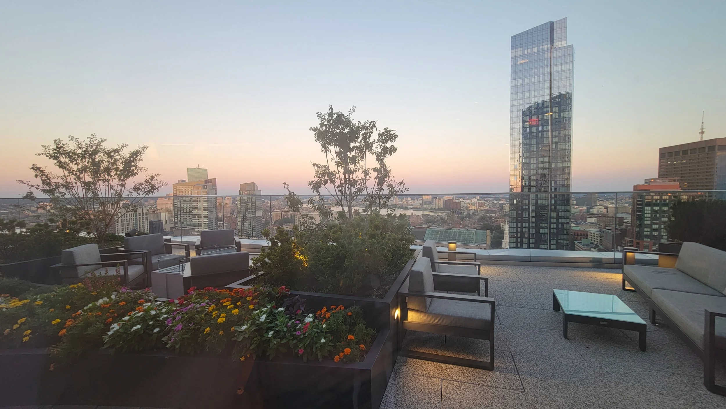 Rooftop patio with seating, potted plants, flowers, and city skyline at sunset.