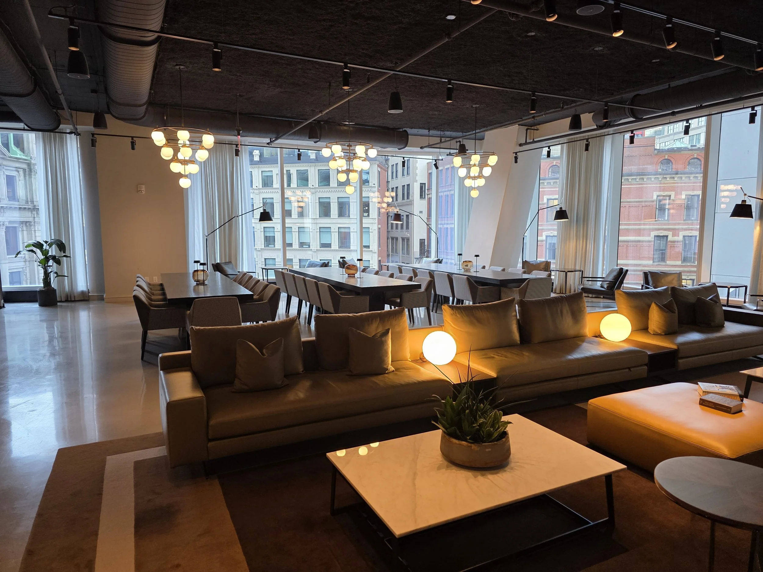 Interior of a modern, stylish lounge or living space with large windows, a long sofa with cushions, a marble coffee table with a plant, and a variety of chairs and tables, illuminated by warm ambient lighting and hanging ceiling lights.