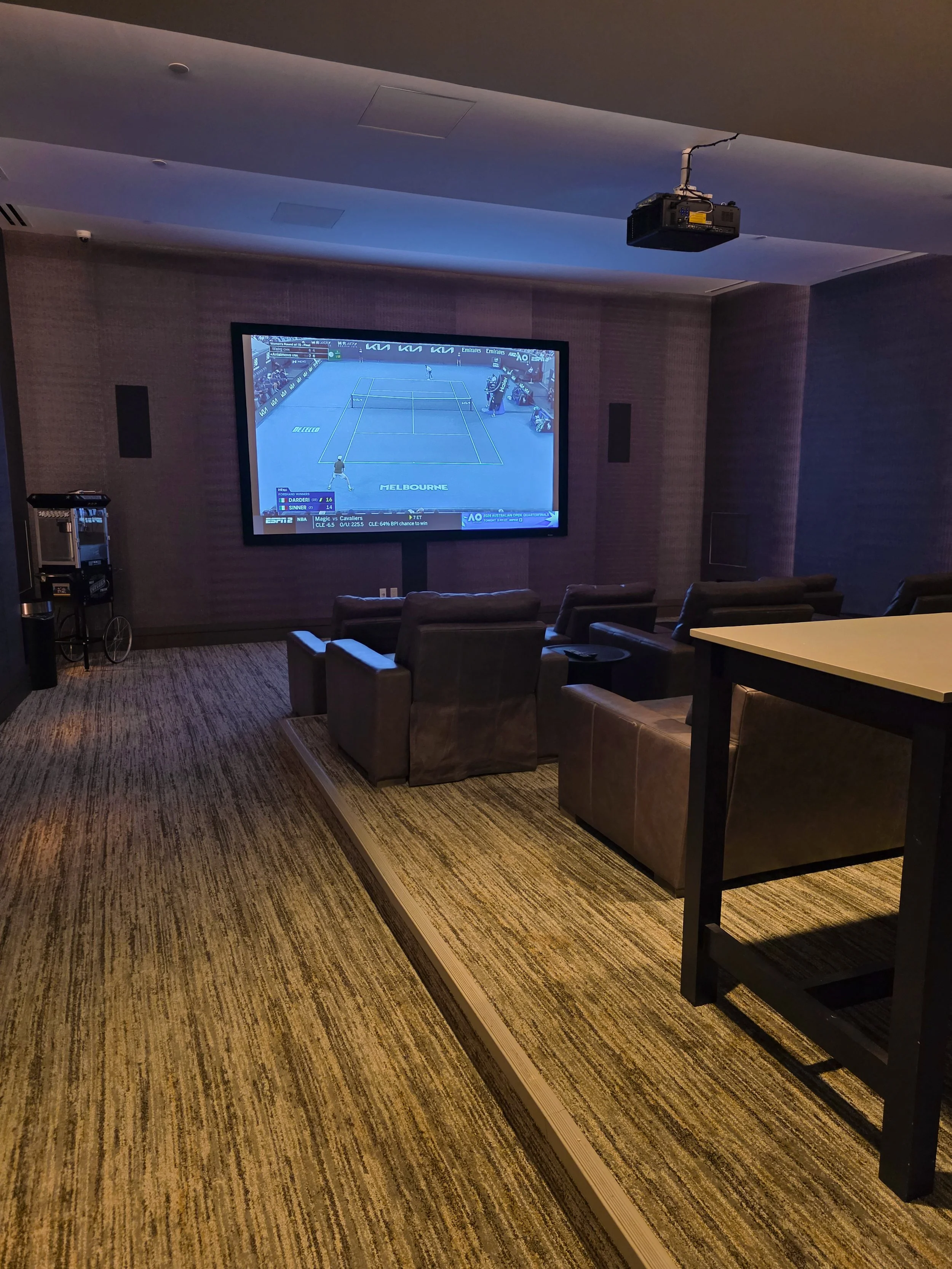 A home theater room with a large screen displaying a tennis match, several recliner chairs, a carpeted floor, and a ceiling-mounted projector.