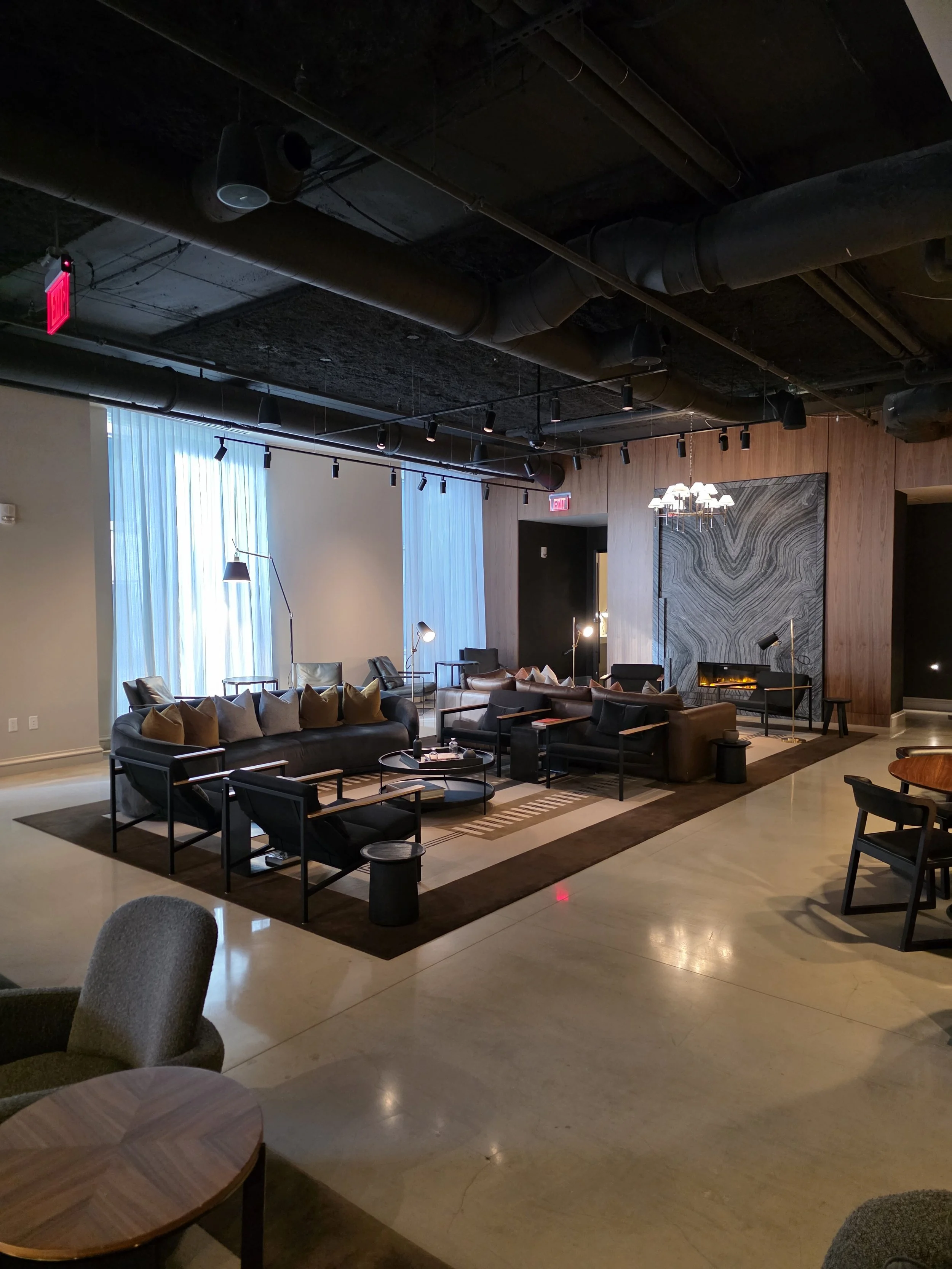 A modern lounge area with black sofas, armchairs, and coffee tables. There are floor and table lamps, large windows with sheer curtains, and a decorative stone wall with a fireplace. The ceiling is black with exposed ductwork and track lighting.