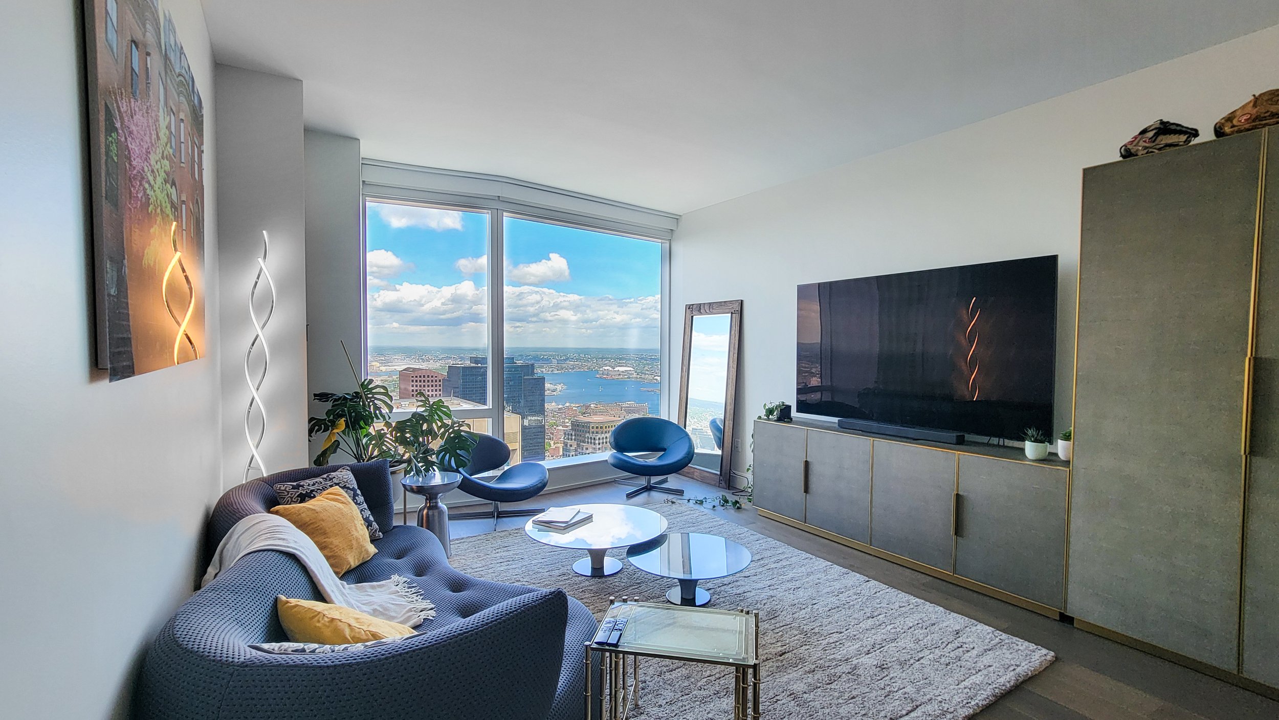 Modern living room with large floor-to-ceiling windows showcasing a city skyline and river view, blue chairs, a textured gray sofa with yellow and white pillows, a glass coffee table, a decorative plant, a tall mirror, and an entertainment center wit