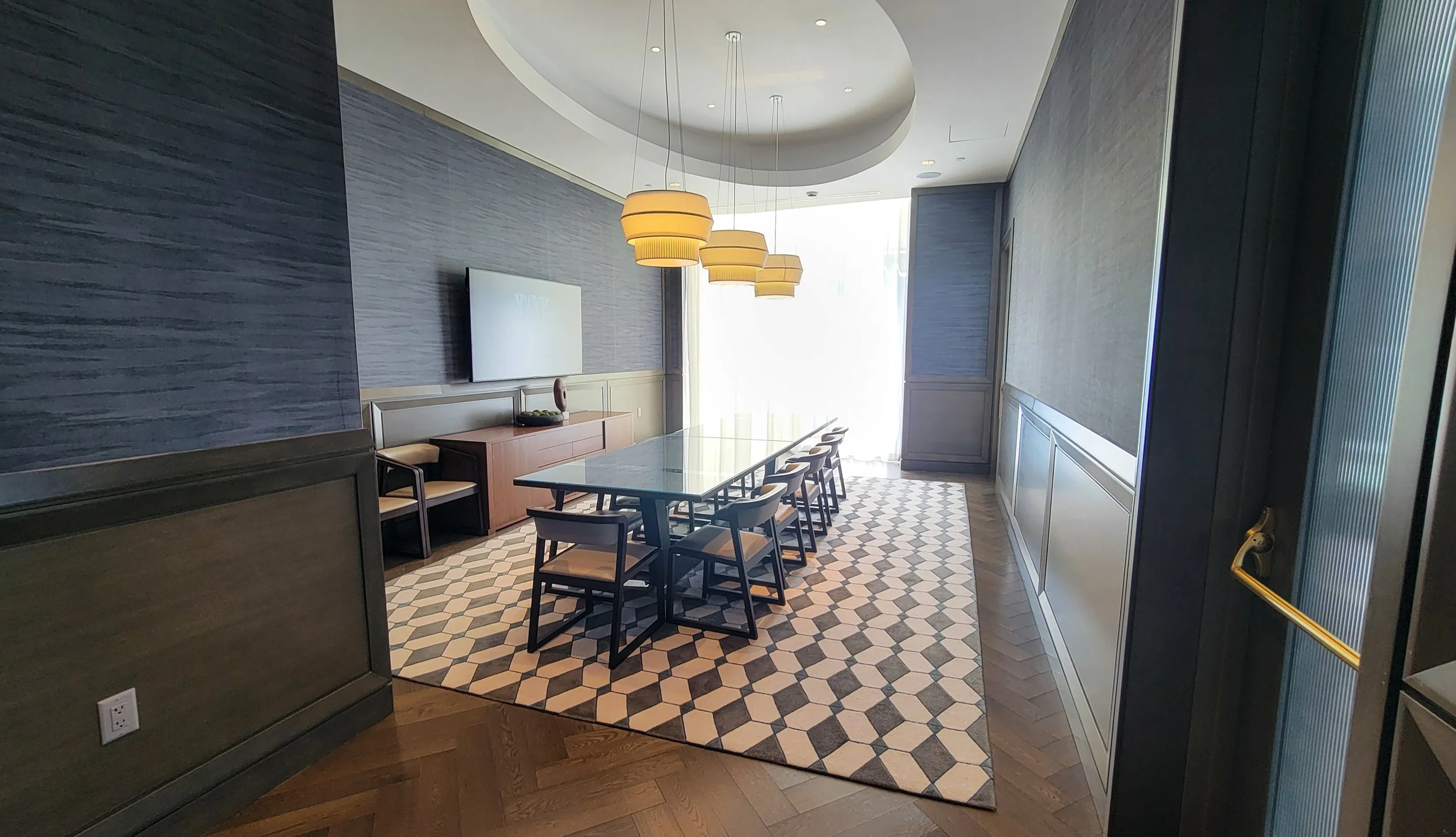 Modern dining conference room with a long table, multiple chairs, a geometric-patterned rug, a wall-mounted TV, and pendant lighting fixtures.