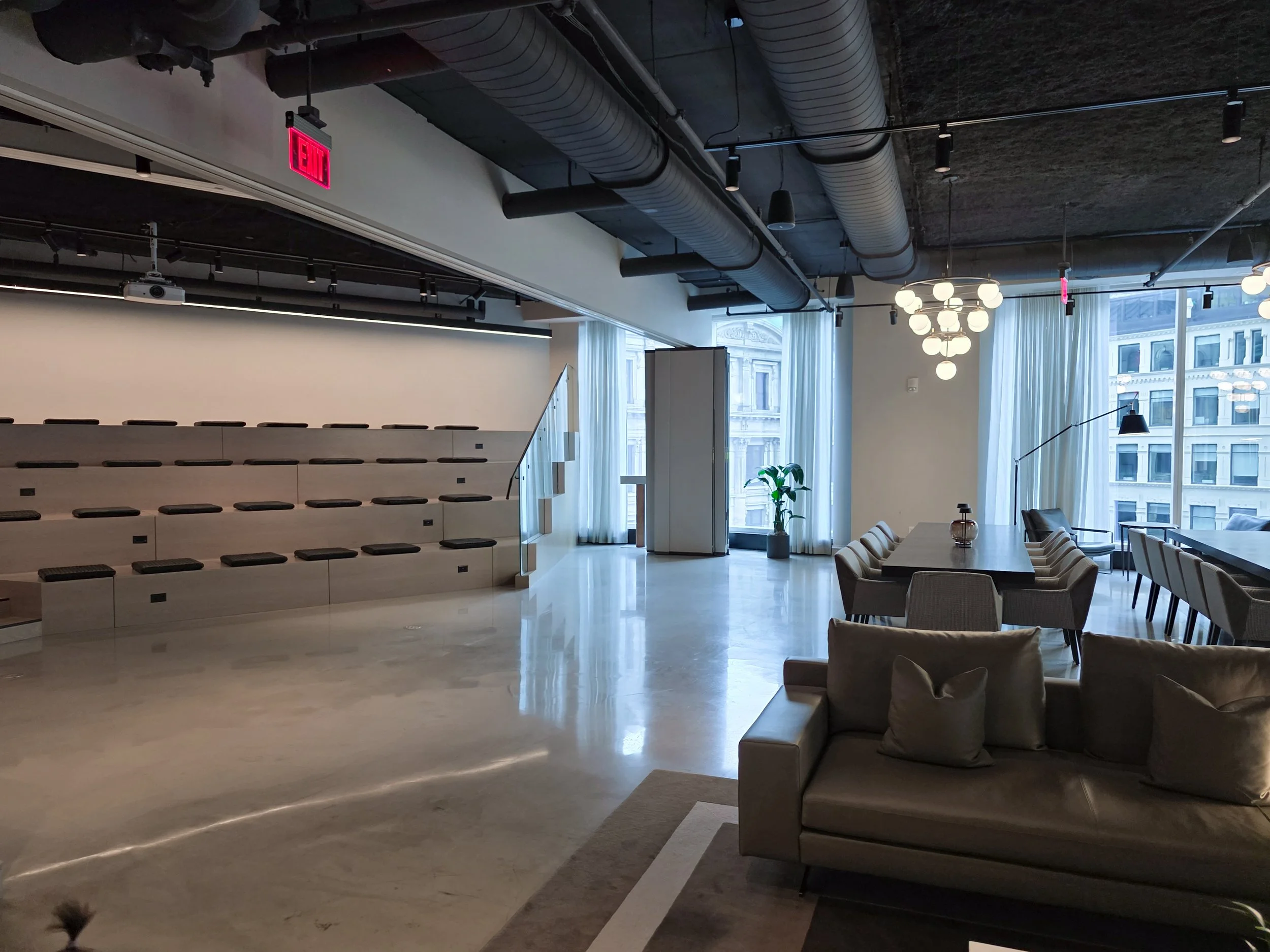 Modern living room or office space with large windows, white curtains, a beige sofa with pillows, dining table with chairs, hanging lights, a potted plant, and exposed ceiling pipes.