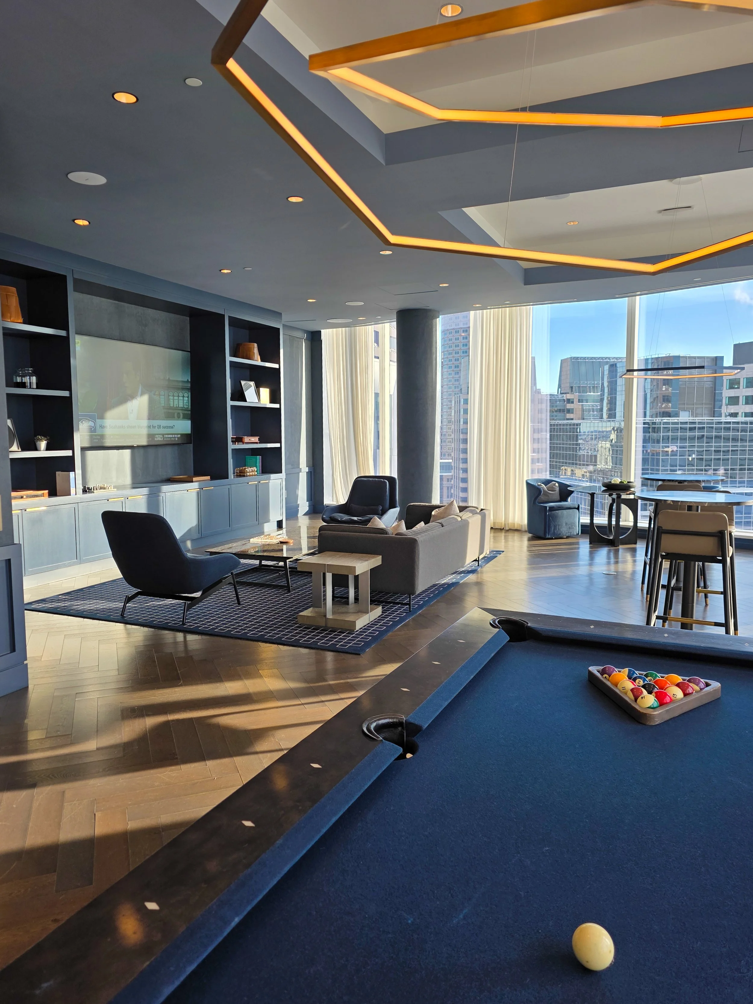 Modern lounge area with large windows letting in natural light, featuring a pool table with a cue ball and rack of balls, a seating area with armchairs and a sofa, a television, and cityscape view.