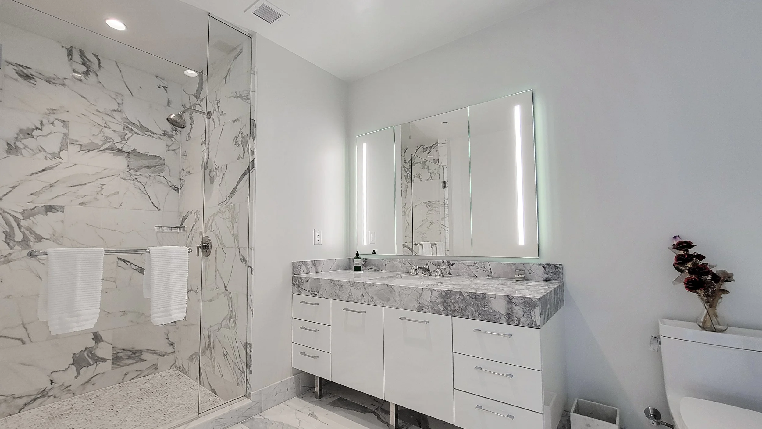 Modern bathroom with a marble-topped vanity, large mirror with light strips, and a walk-in shower with marble walls and two white towels on a rail.
