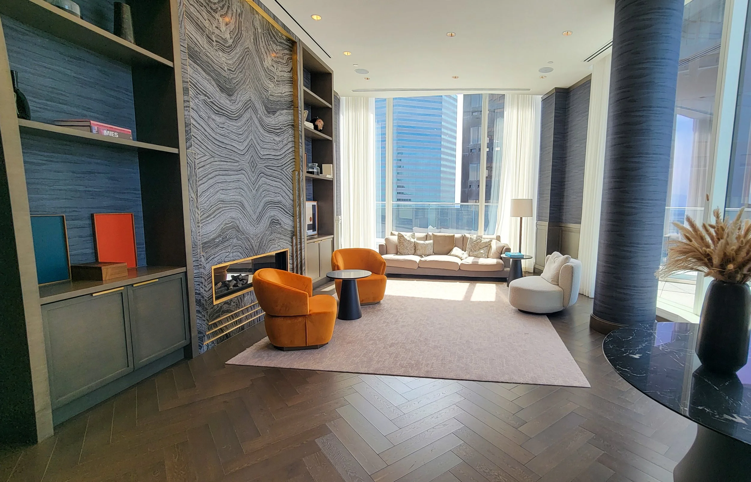 A modern living room with large windows, a beige couch, two orange chairs, a white chair, a small round table, a black side table, a lamp, and a decorative vase with dried plants. The walls have textured blue-gray wallpaper, and a bookshelf is built 