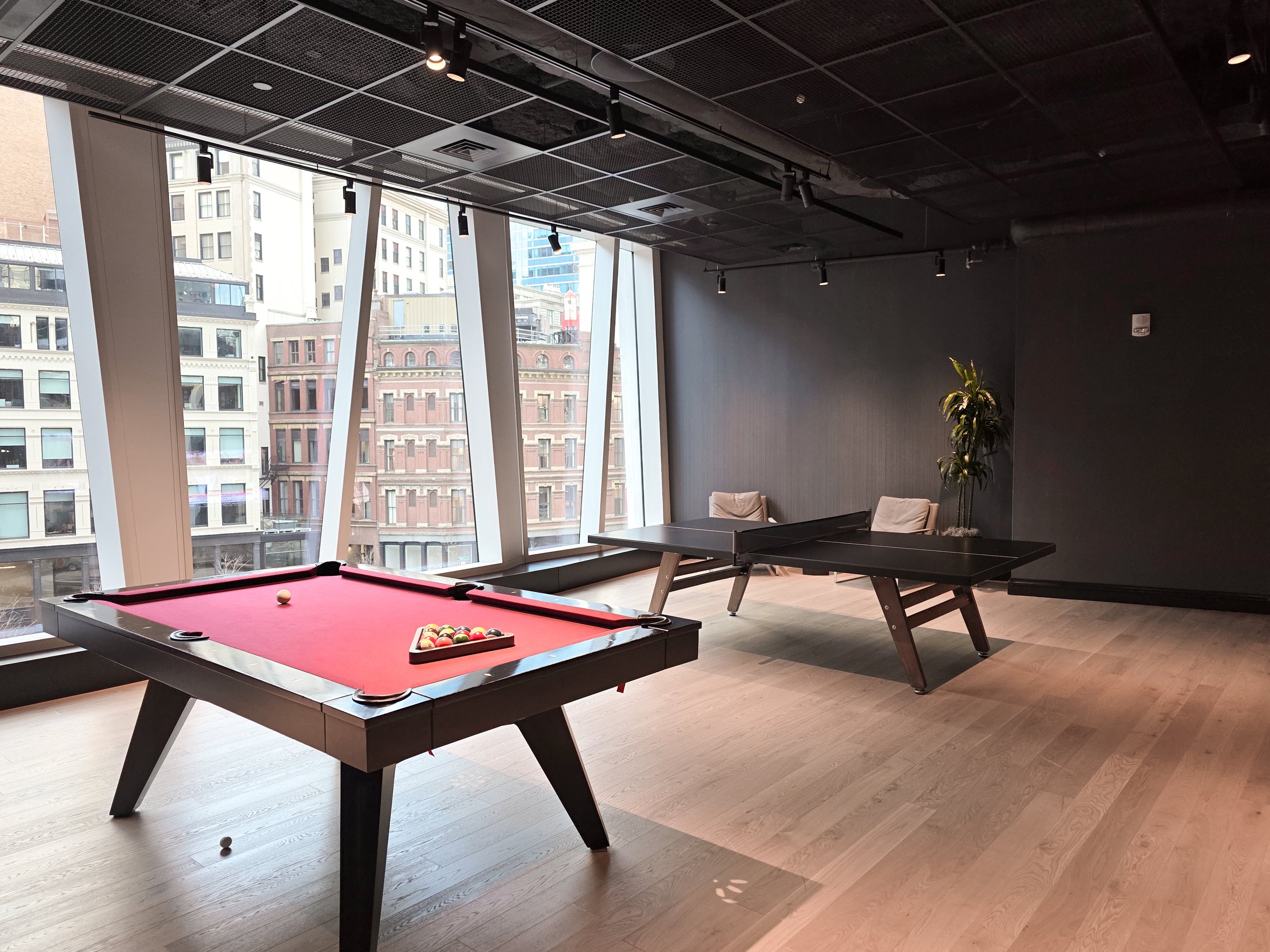 An indoor recreation area with a pool table and a ping pong table, in a room with large windows showing city buildings outside.