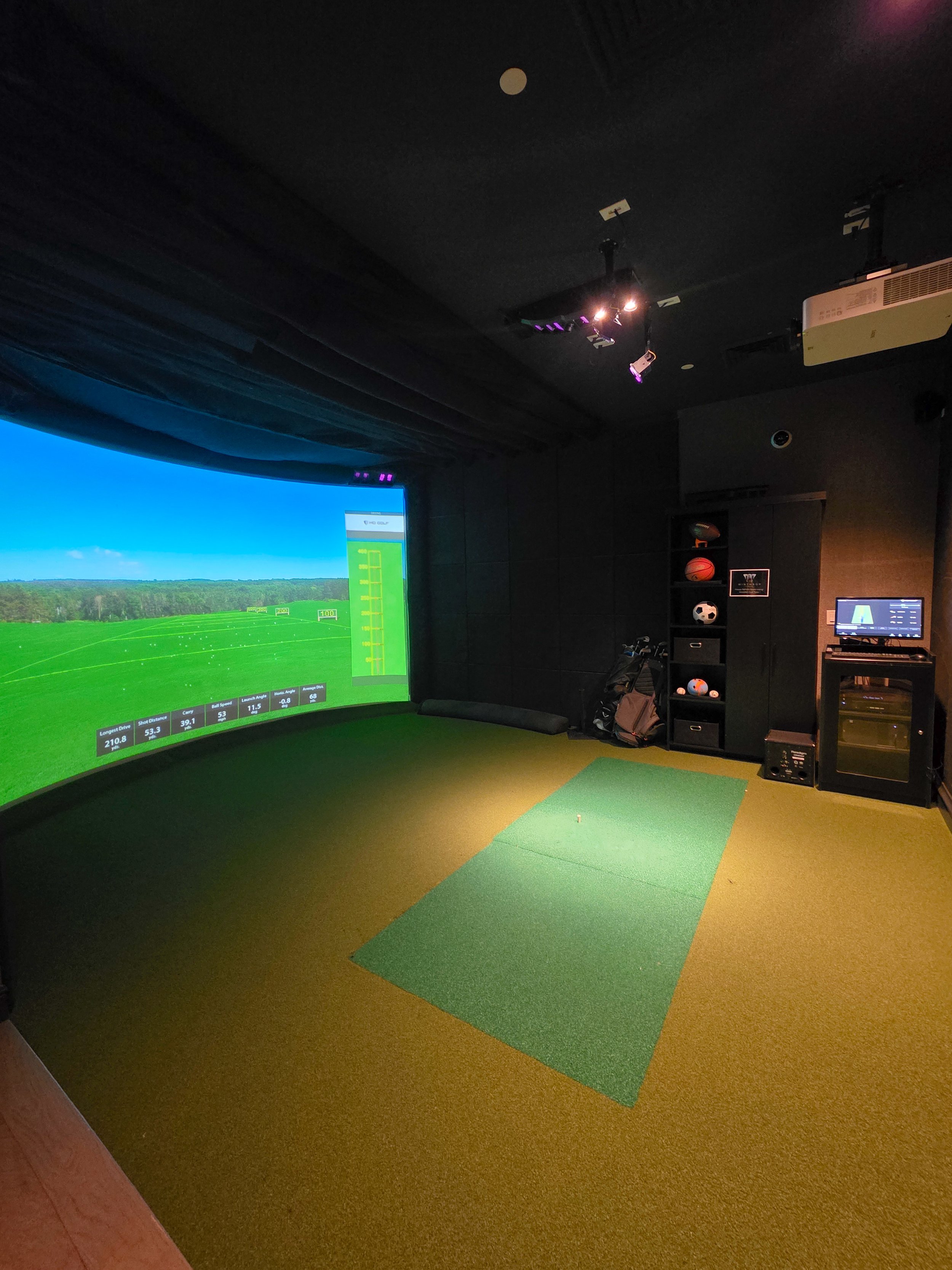 Indoor golf simulator room with a large screen displaying a virtual golf course, artificial turf mat, and golf clubs in a black shelving unit.