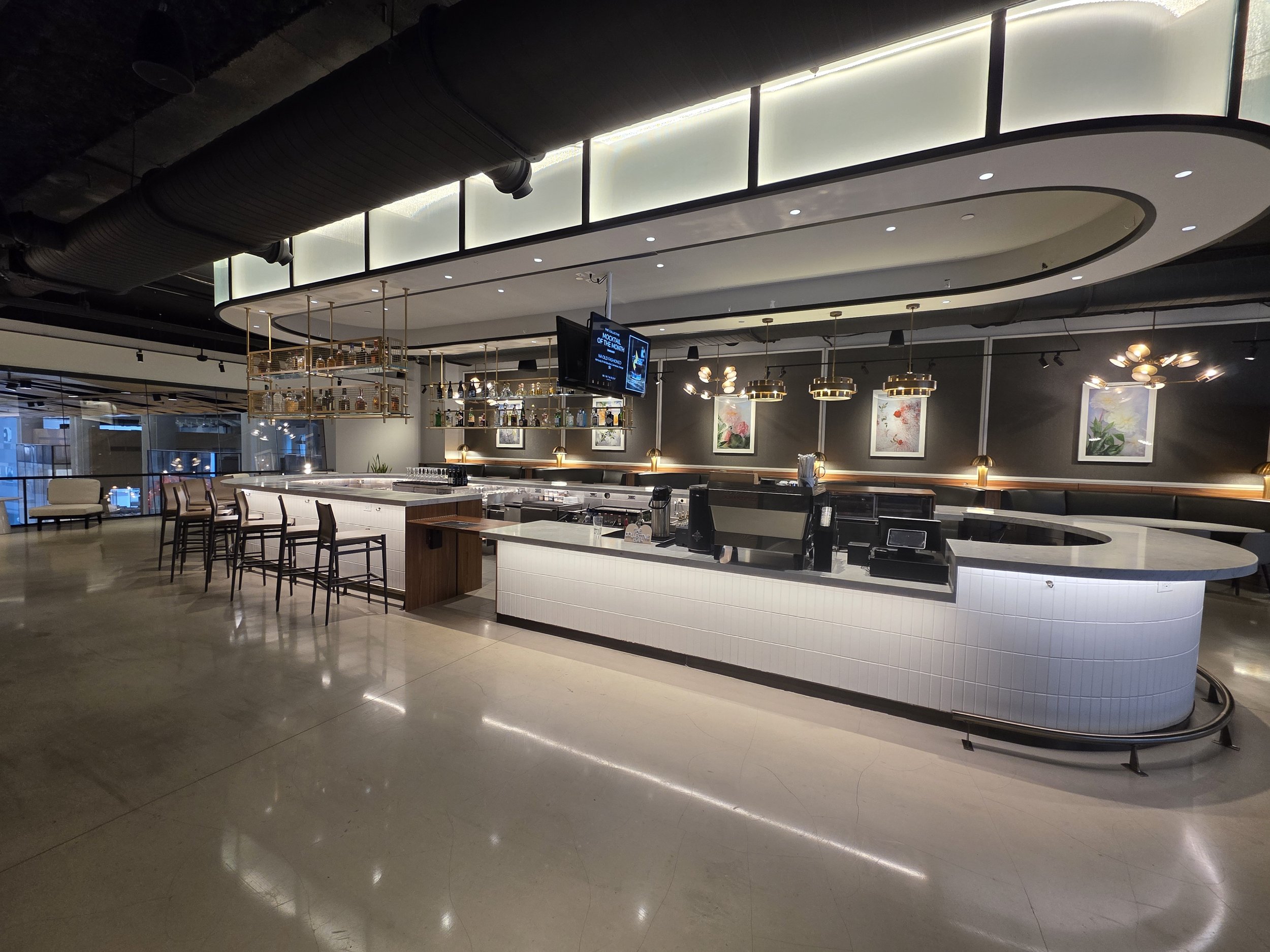 Modern, stylish bar with white curved counter, bar stools, industrial ceiling ducts, artwork on dark wall, and hanging pendant lights.