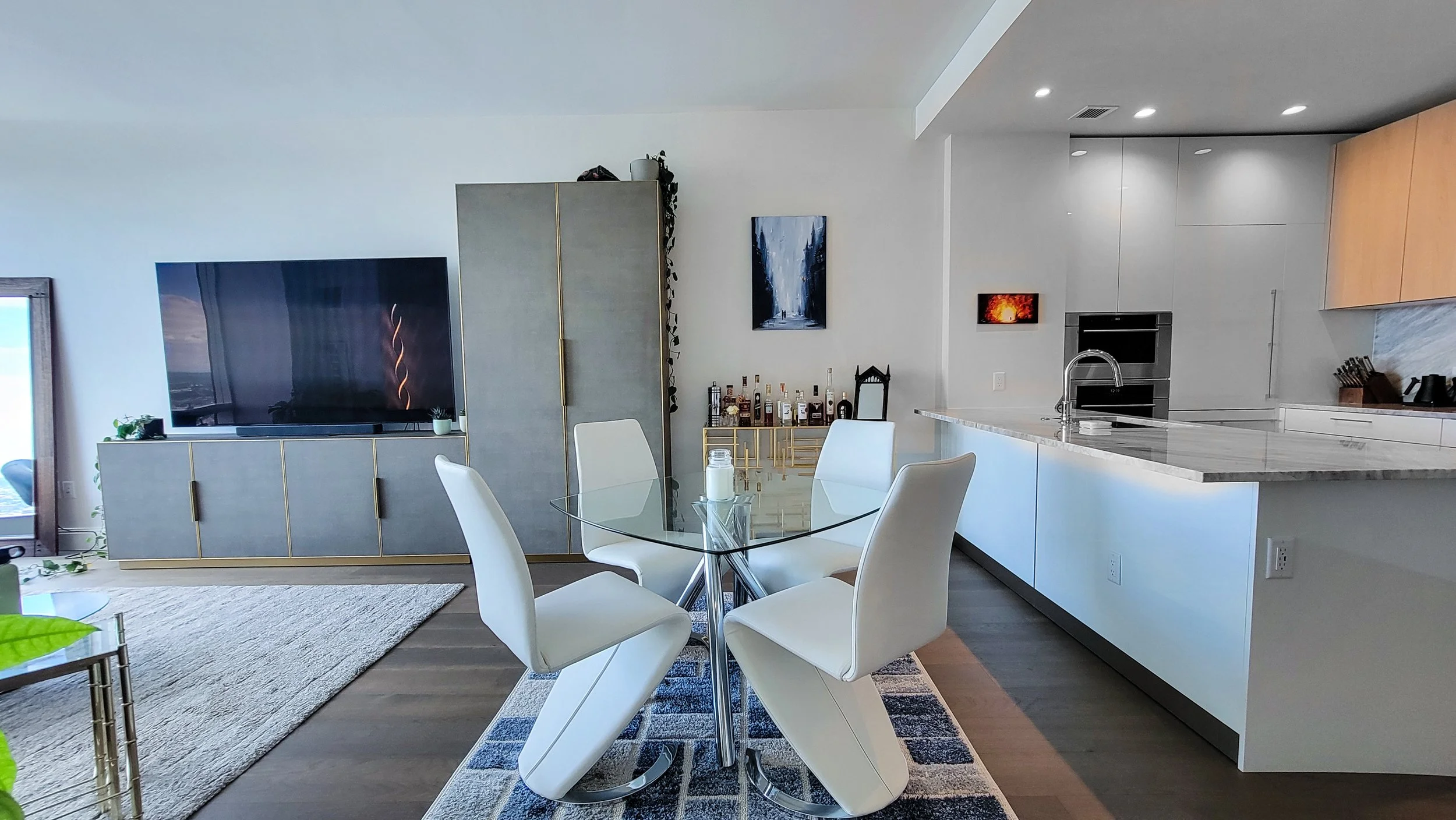Modern living room and kitchen with a glass dining table, white chairs, gray cabinet with TV, bar area with liquor bottles, and minimalist decor.