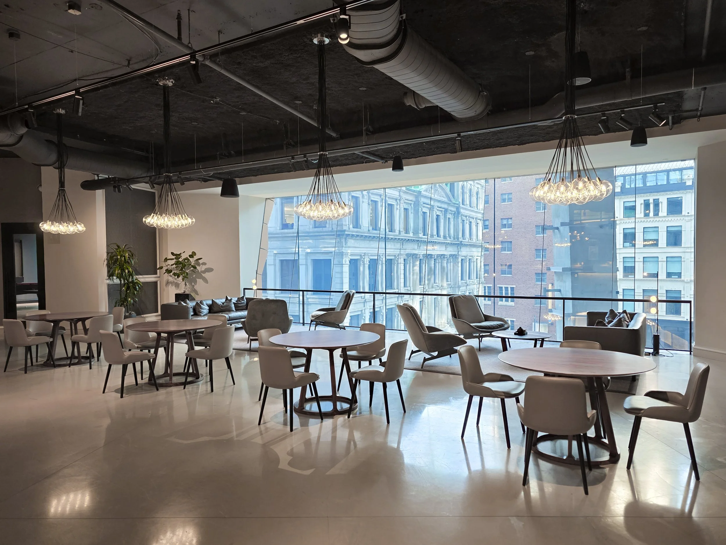 Modern lounge with round and rectangular tables, beige chairs, gray armchairs, plants, and large windows overlooking city buildings