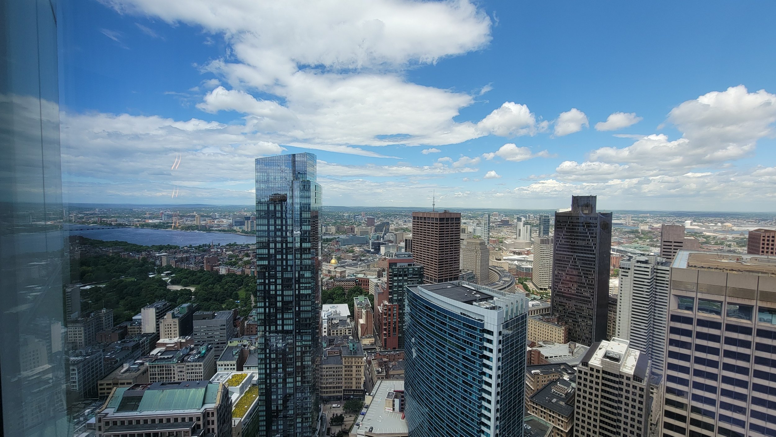 A panoramic view of a city skyline with tall modern skyscrapers, a river, and a partly cloudy blue sky.