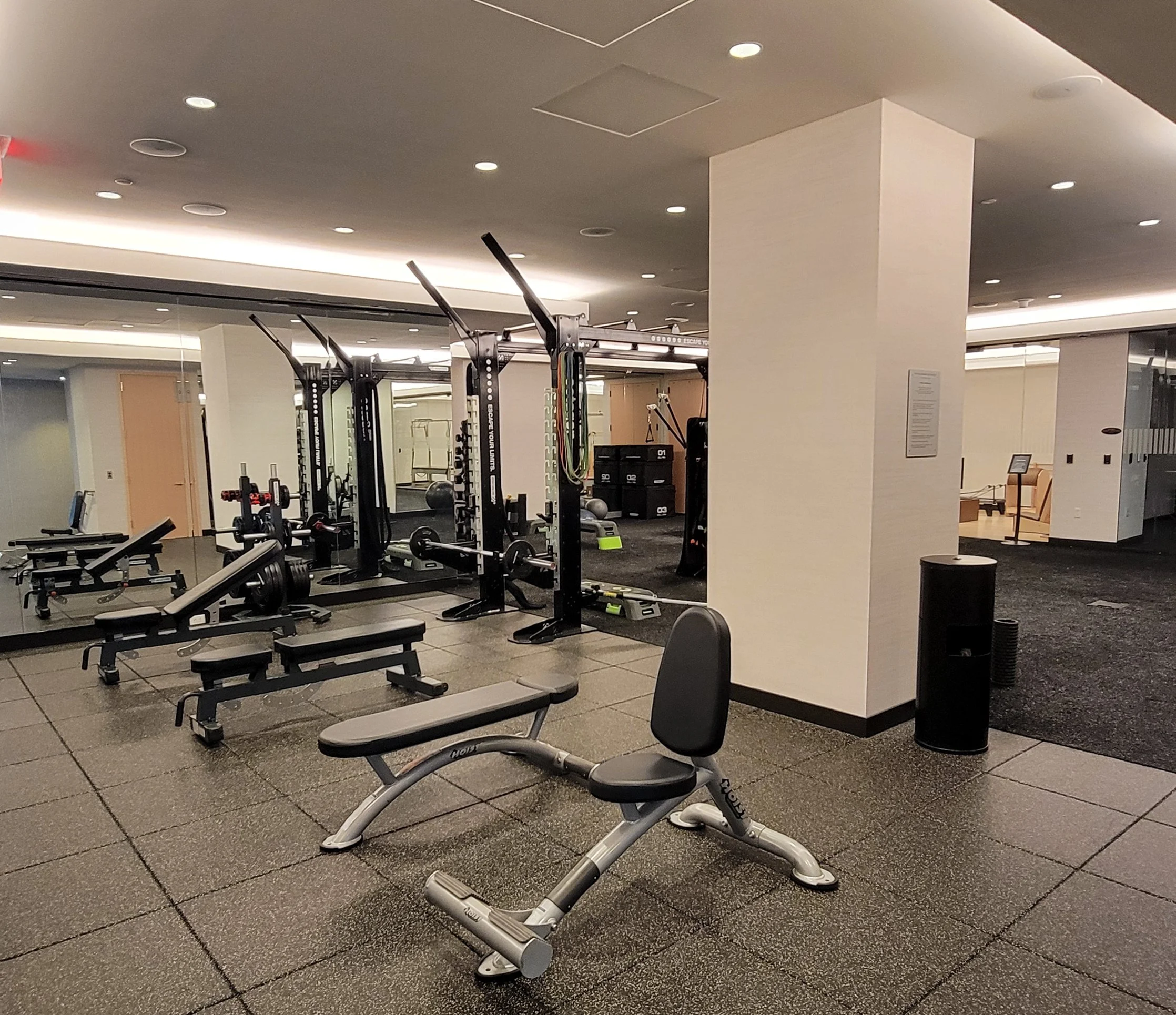 Empty gym with weightlifting equipment, benches, a mirror, and a white wall with a column in a modern fitness center.