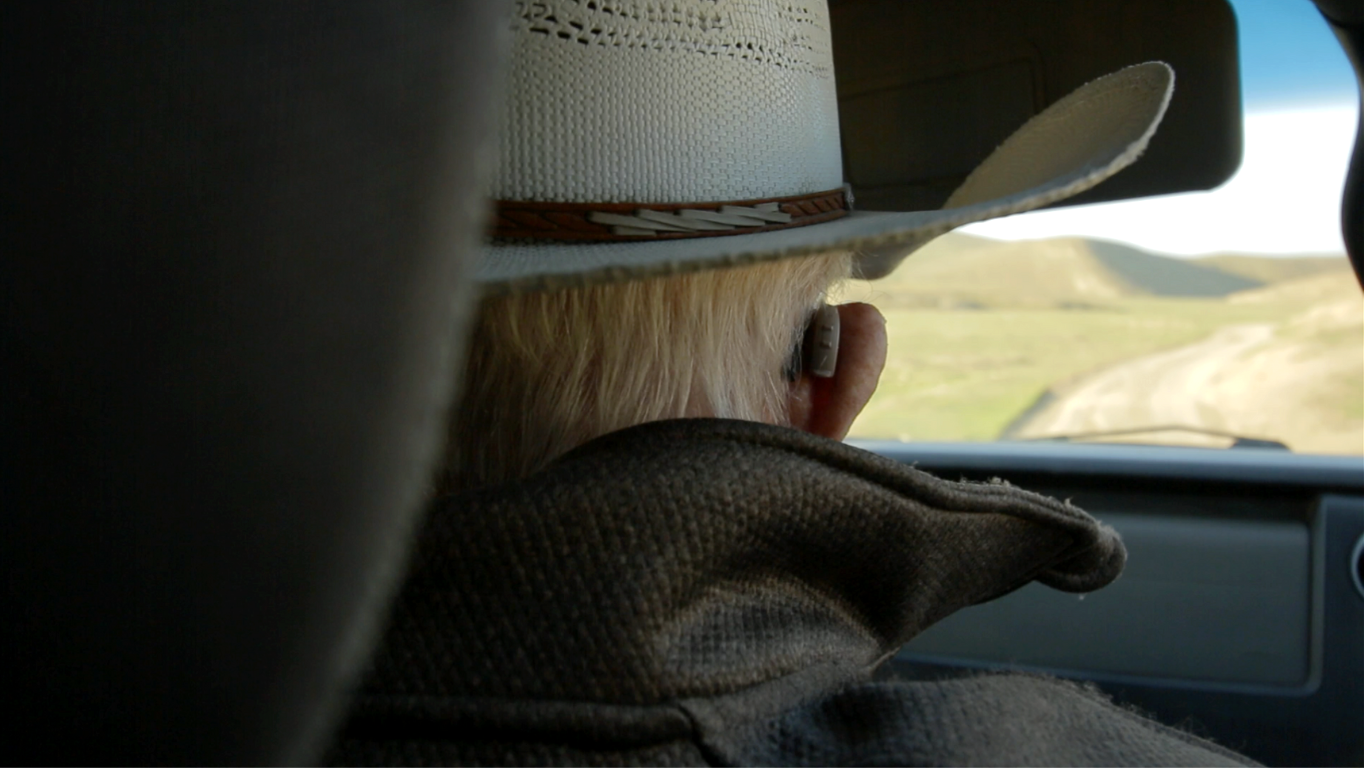 Person with blonde hair wearing a cowboy hat, seated in a vehicle, looking out the front windshield at a rural landscape with rolling hills.