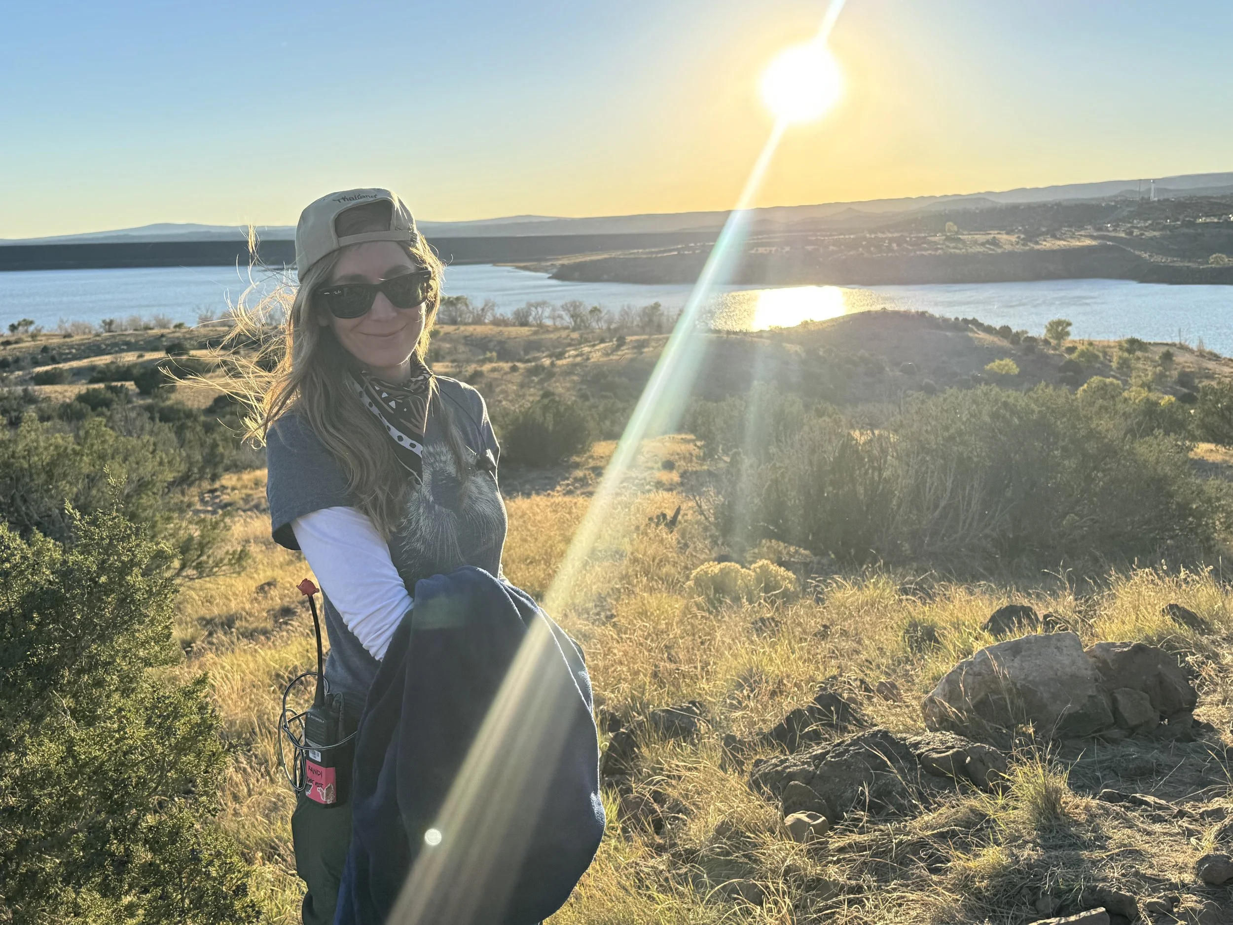 Woman wearing sunglasses, a cap, and a bandana around her neck, smiling outdoor during sunset near a lake with hills in the background.