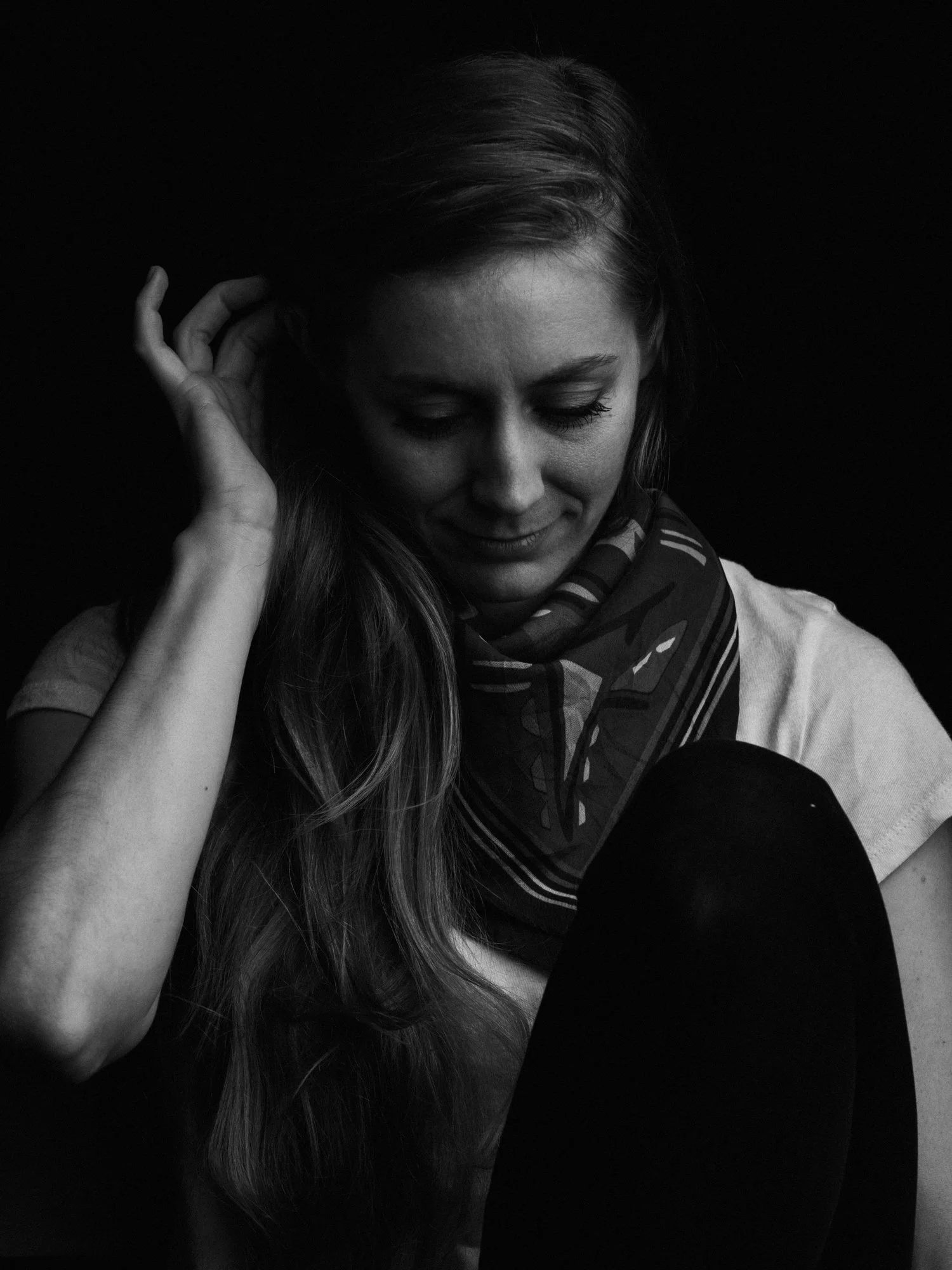 Black and white photo of a woman with long hair, wearing a scarf, smiling gently with eyes closed, in a relaxed pose against a dark background.