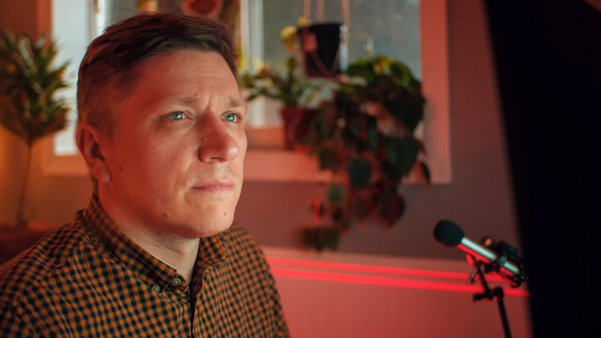 A man with blue eyes and short brown hair looking to his left indoors, with a window and potted plant in the background. There is a microphone on a stand in front of him.