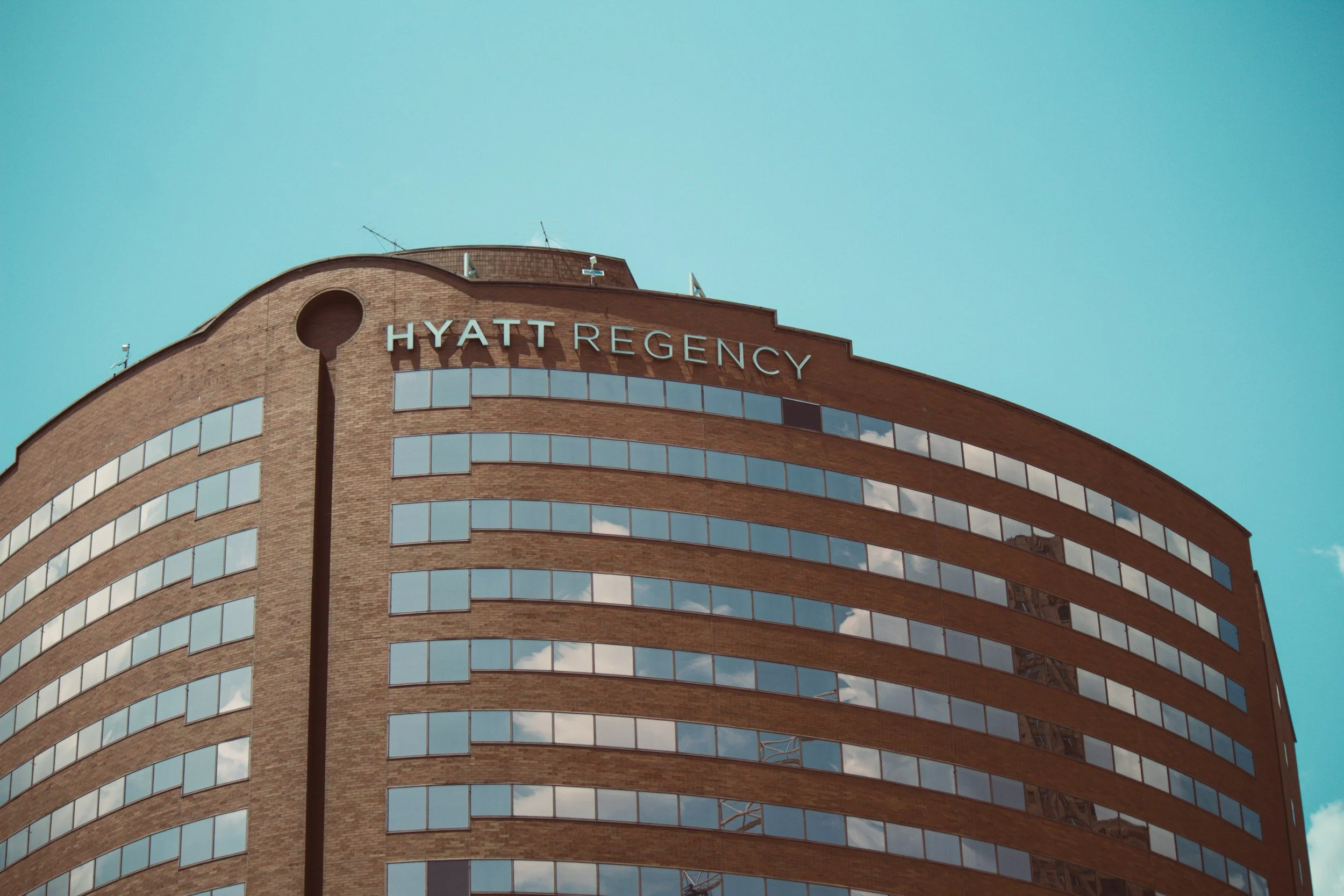 Top portion of a curved brick hotel building with large blue glass windows and the words 'Hyatt Regency' on the upper part, with blue sky in the background.
